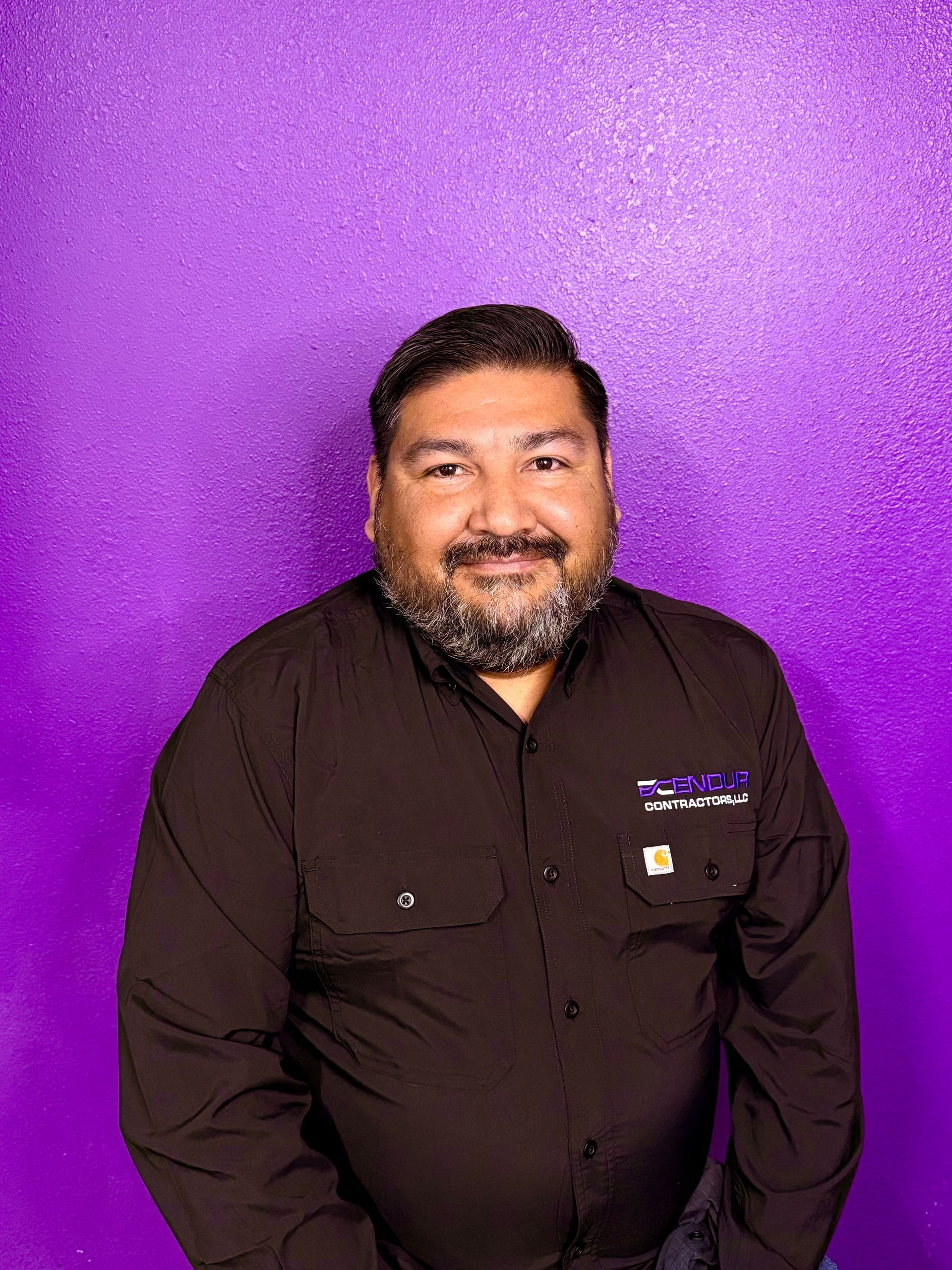 Bald man wearing a black shirt, standing in front of a purple wall.