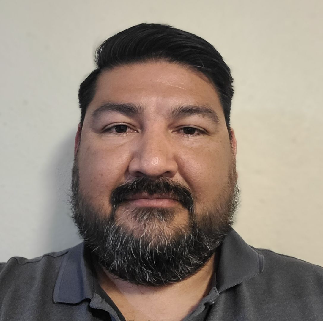 Man with dark hair and beard, wearing a gray shirt, looking at the camera.