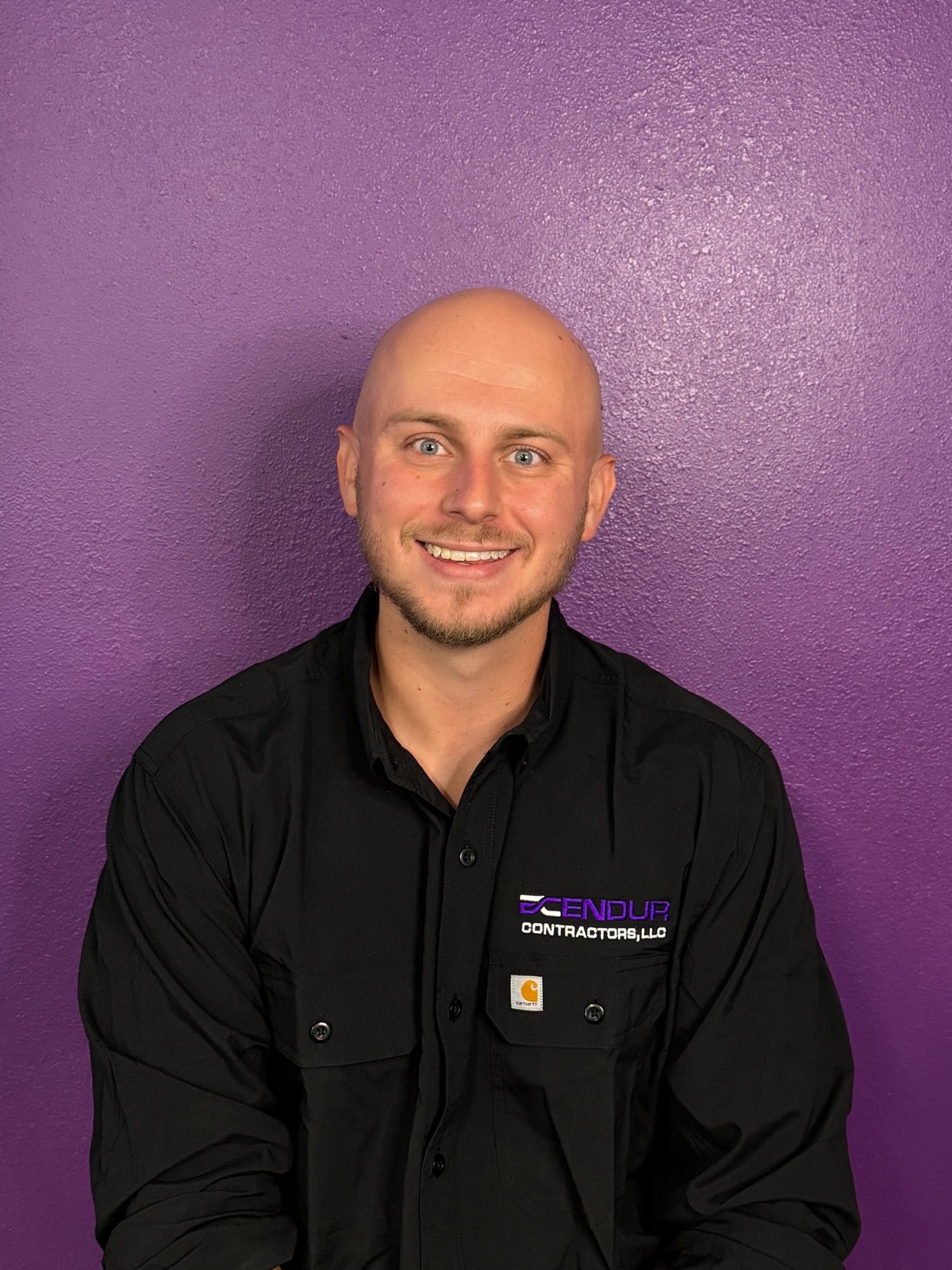 Smiling bald man in a black shirt standing against a purple wall