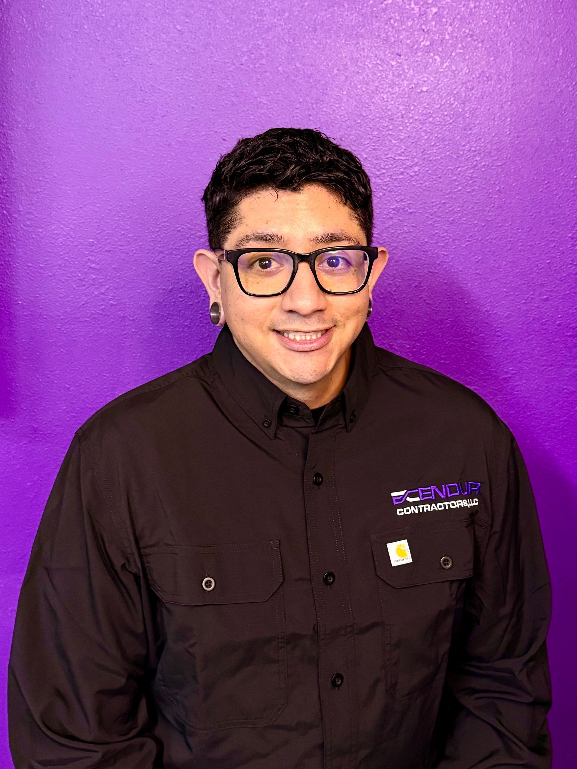 Man in black shirt smiling in front of a purple wall. The shirt has a logo.