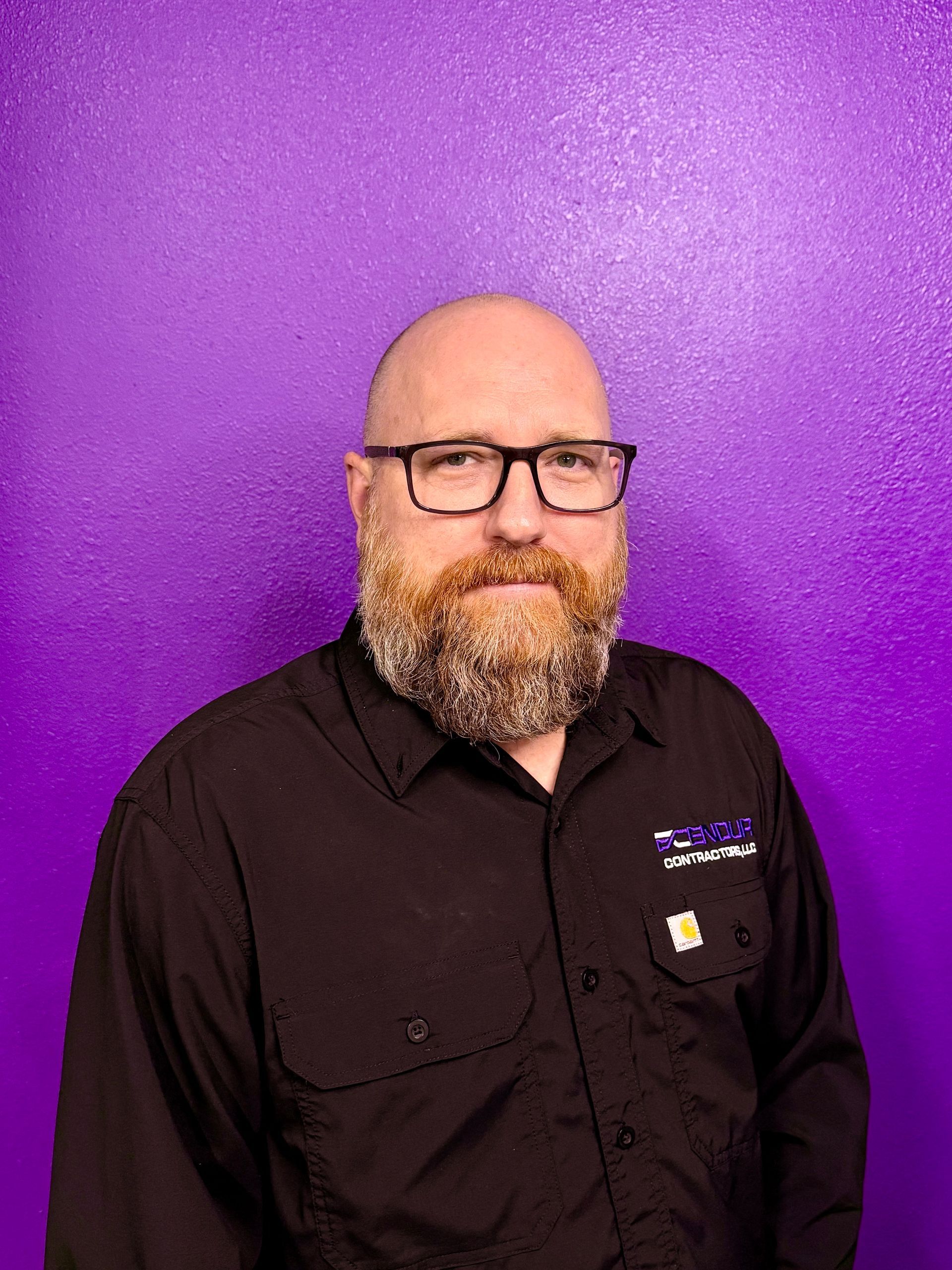 Bald man with glasses and reddish beard, black shirt, standing in front of a purple wall.