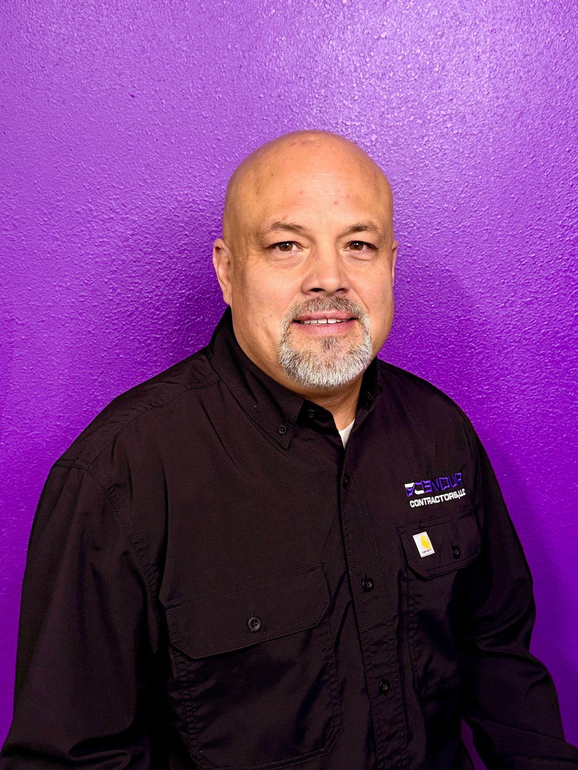 Bald man with a beard, wearing a black shirt, smiles in front of a purple background.