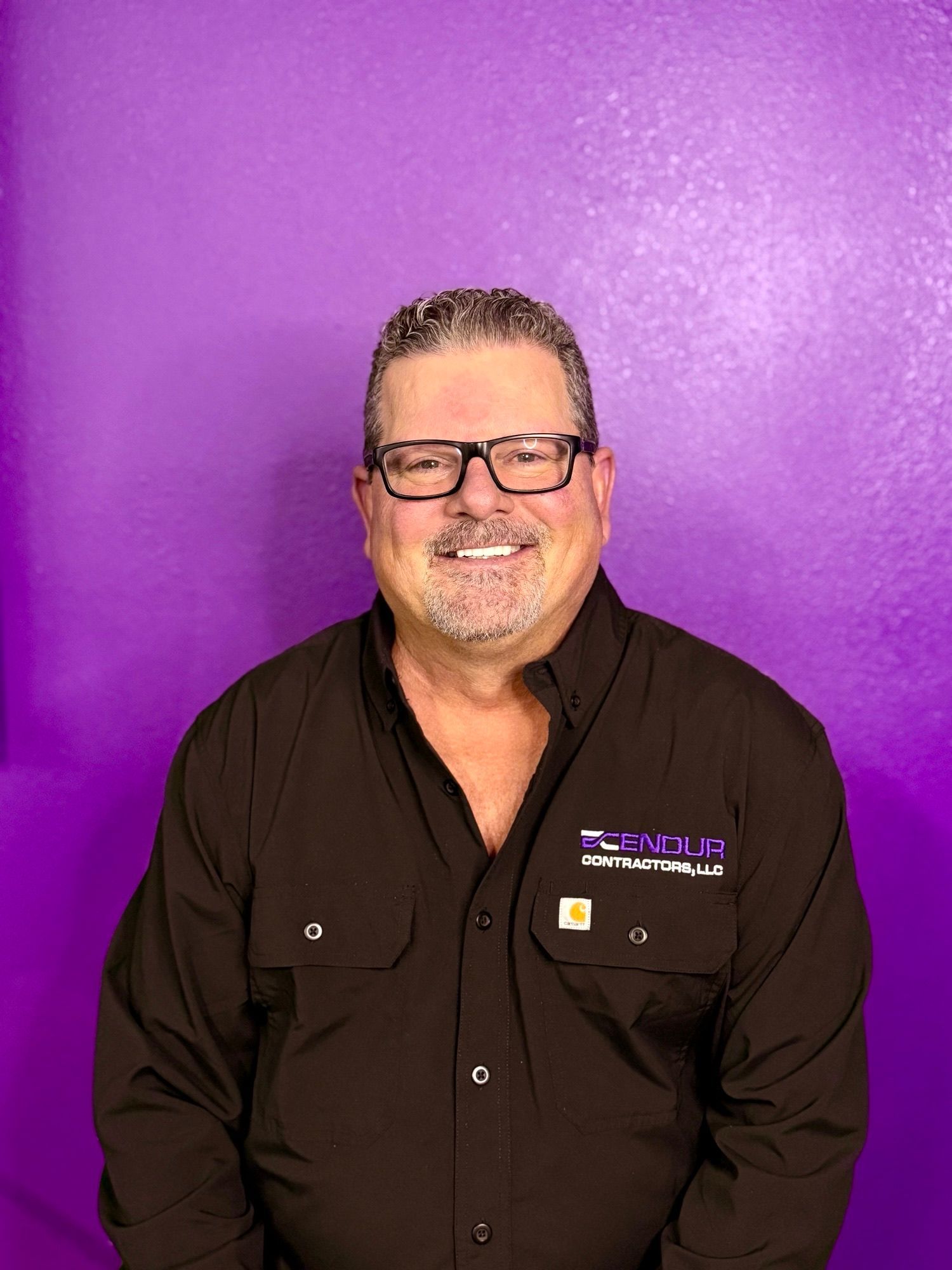 Smiling person in a black shirt with glasses, standing against a purple background.