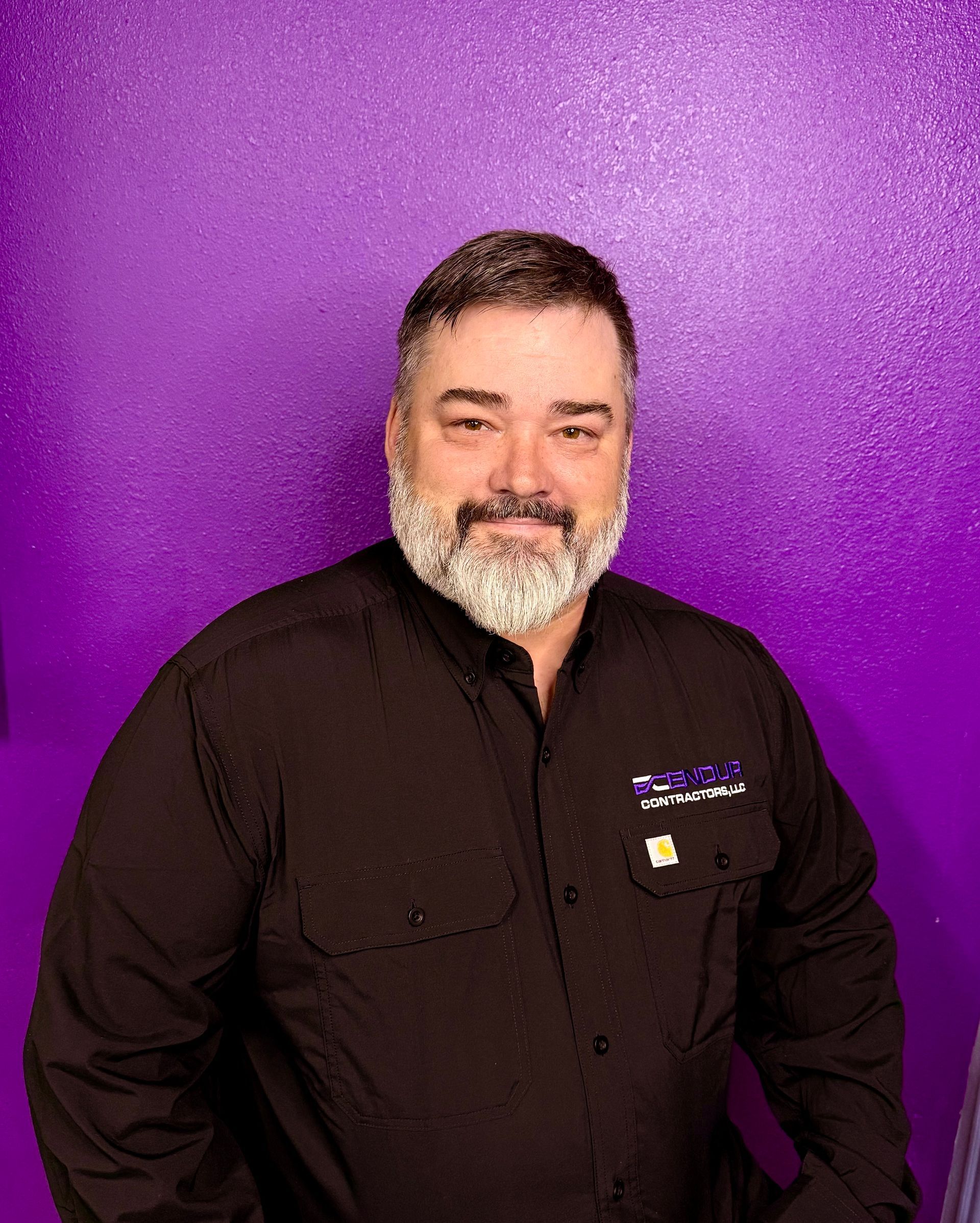 Man with gray beard in black shirt smiling in front of a purple wall.