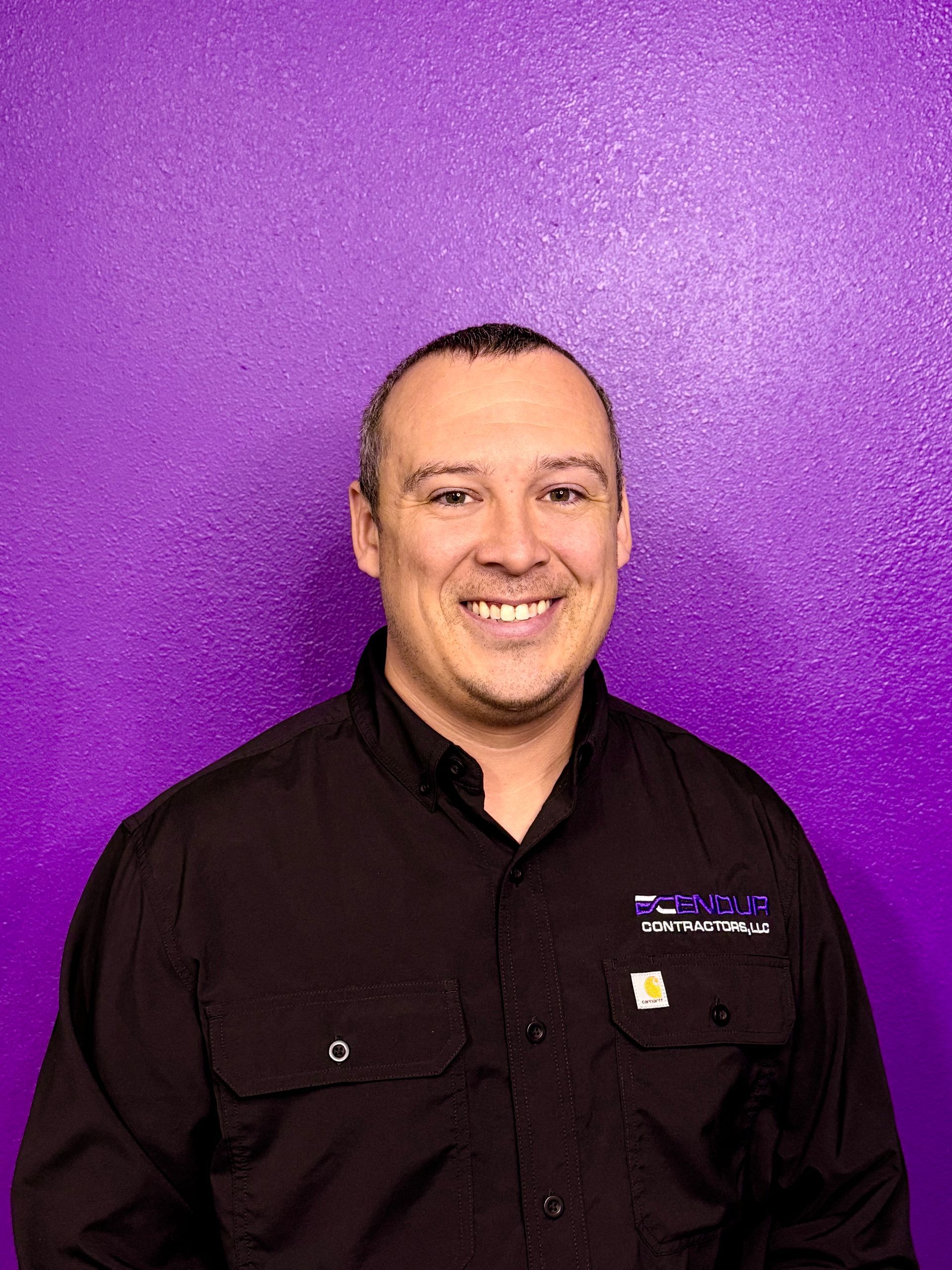 Bald man wearing a black shirt, standing in front of a purple wall.