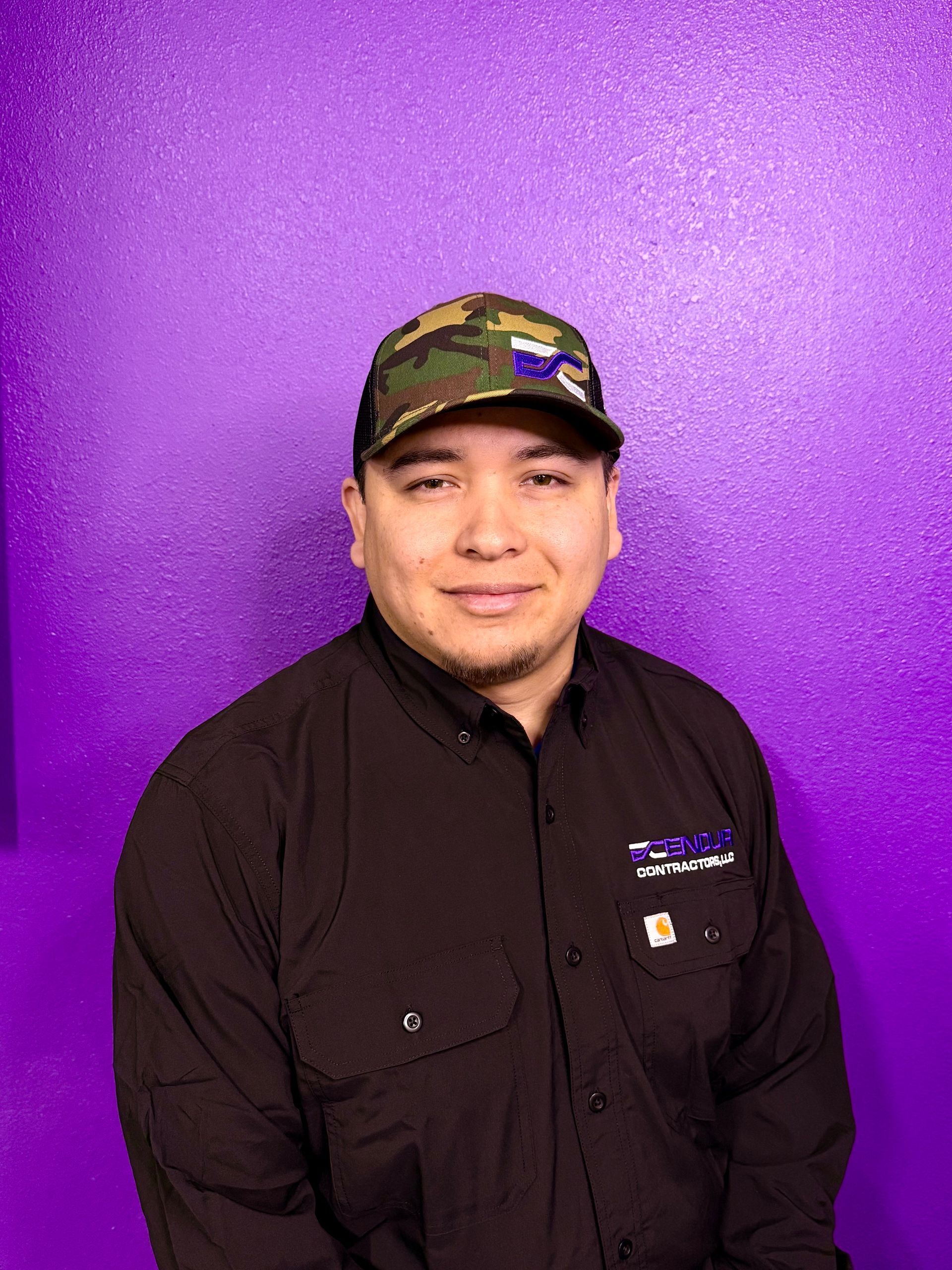 Man in camo cap and black shirt smiles against a purple wall.