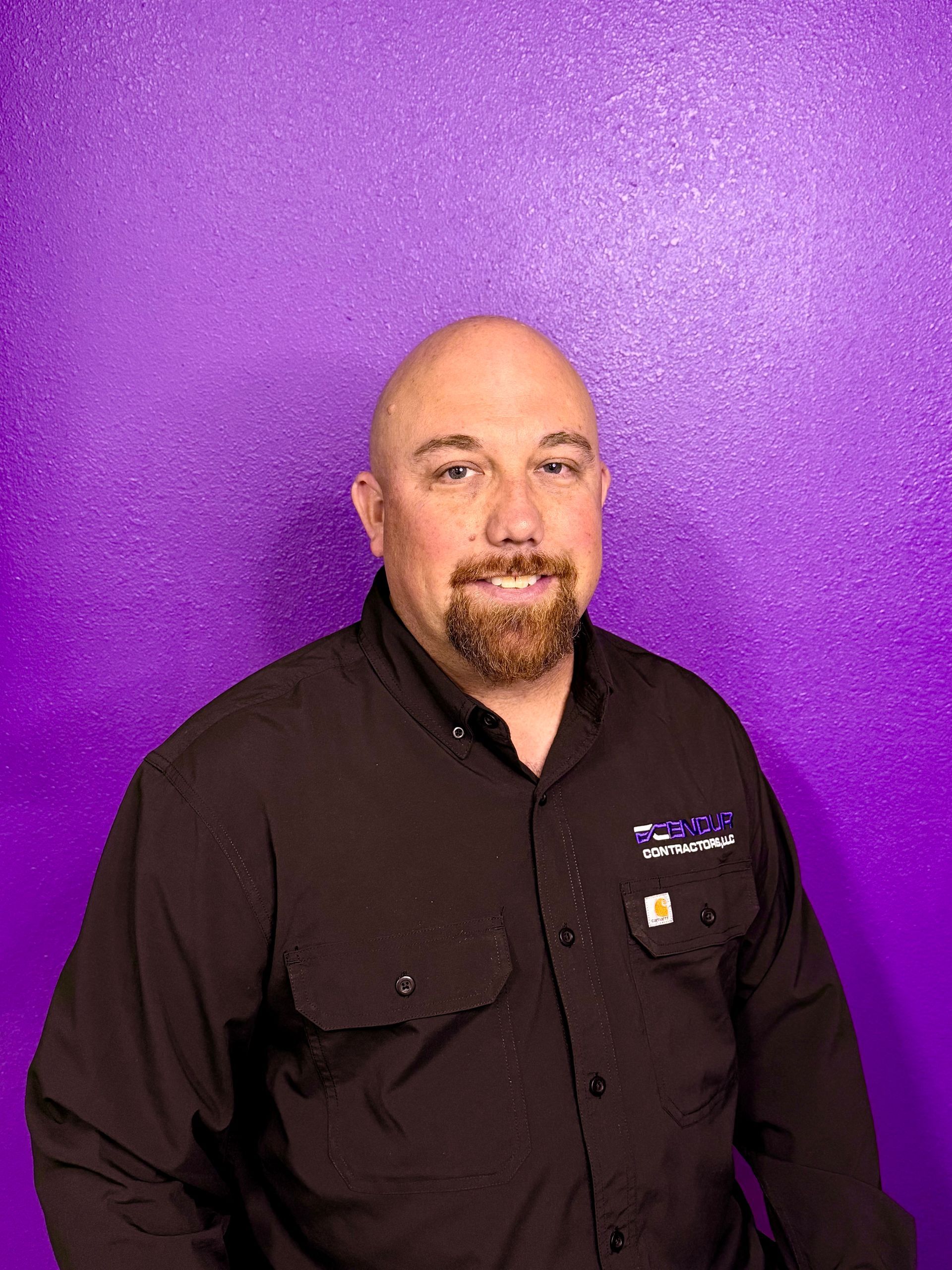 Man with a bald head and goatee smiles, wearing a black shirt, against a purple textured background.