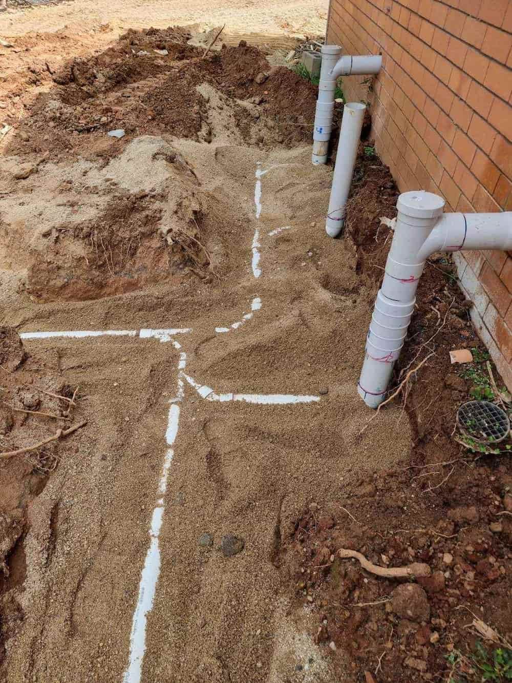 White PVC Pipes Laid in a Trench Along a Brick Wall. the Ground is Covered in Sand — D & B Plumbing NQ in Smithfield, QLD