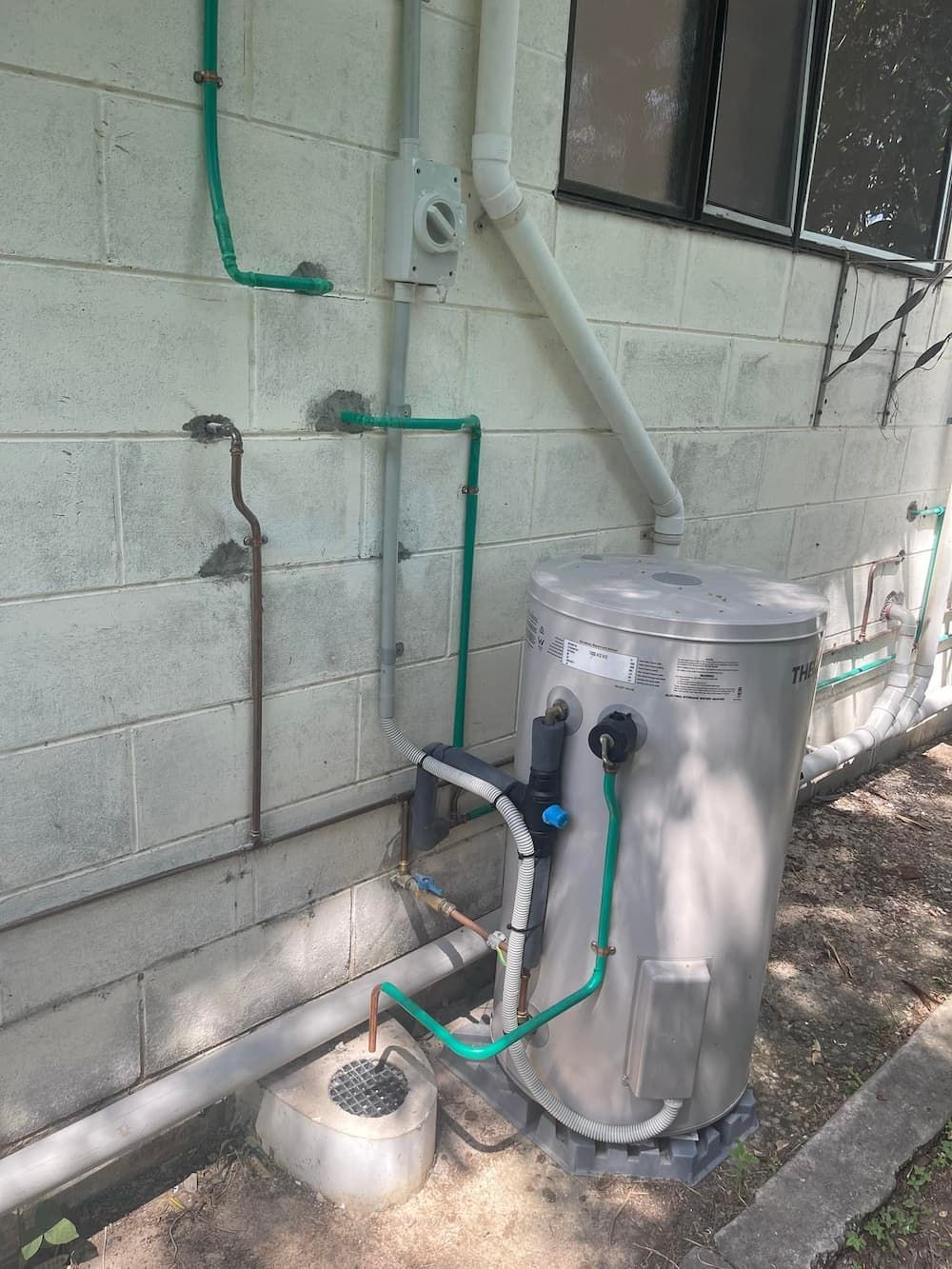 A Water Heater is Installed Outside Against a Cinder Block Wall, With Green Pipes — D & B Plumbing NQ in Trinity Beach, QLD