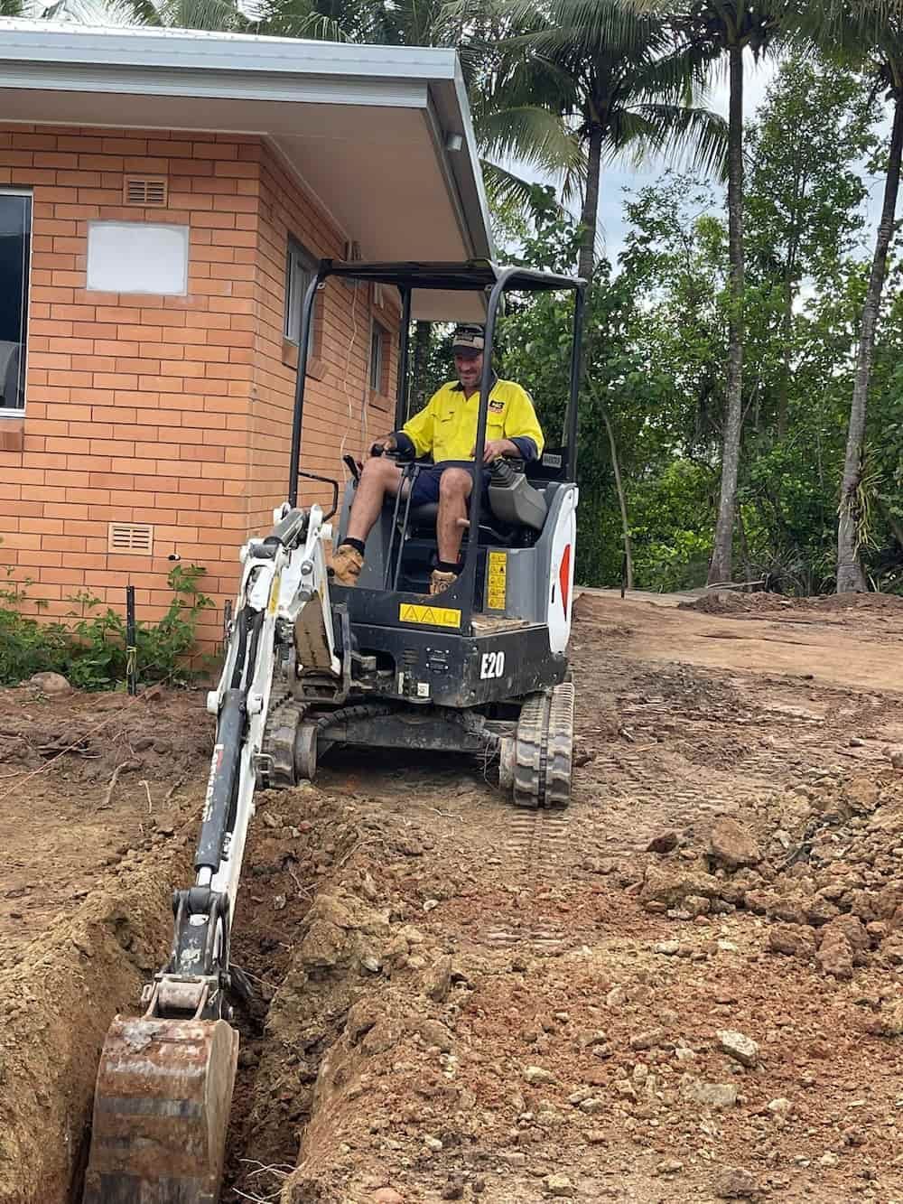 A Person Operates a Small Excavator, Digging a Trench Near a Brick Building — D & B Plumbing NQ in Trinity Beach, QLD