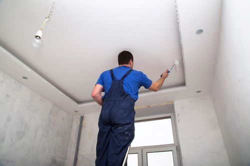 A man is standing on a ladder painting the ceiling of a room.