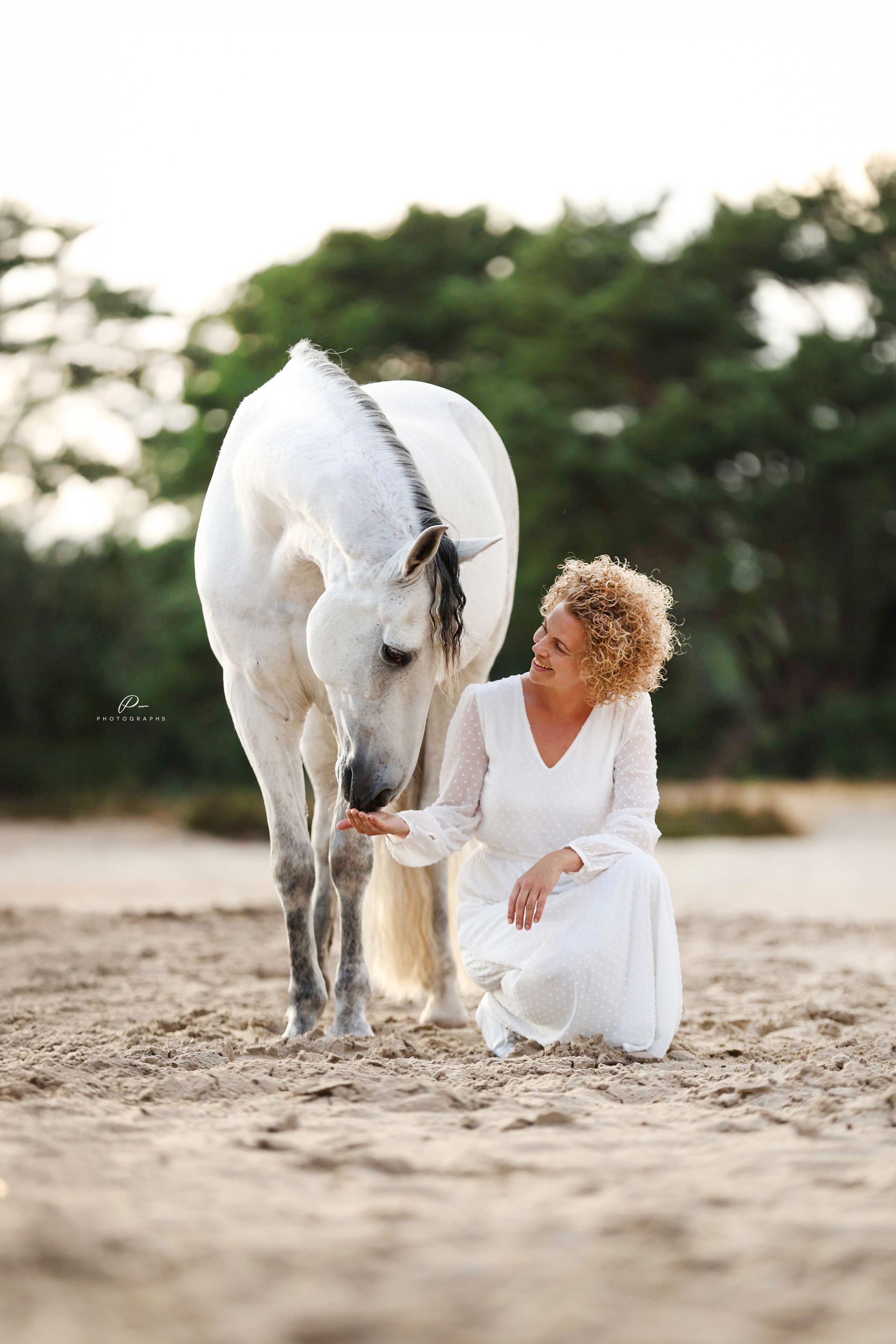 Vrouw in witte jurk knielt en aait een grijs paard op een zanderige ondergrond, met bomen op de achtergrond.