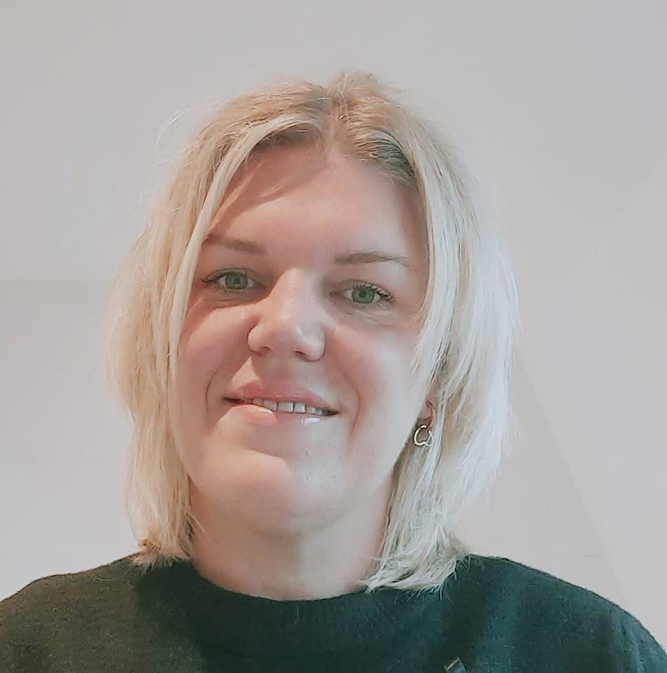 Blonde vrouw, gekleed in een zwarte trui, lacht en staat binnen.