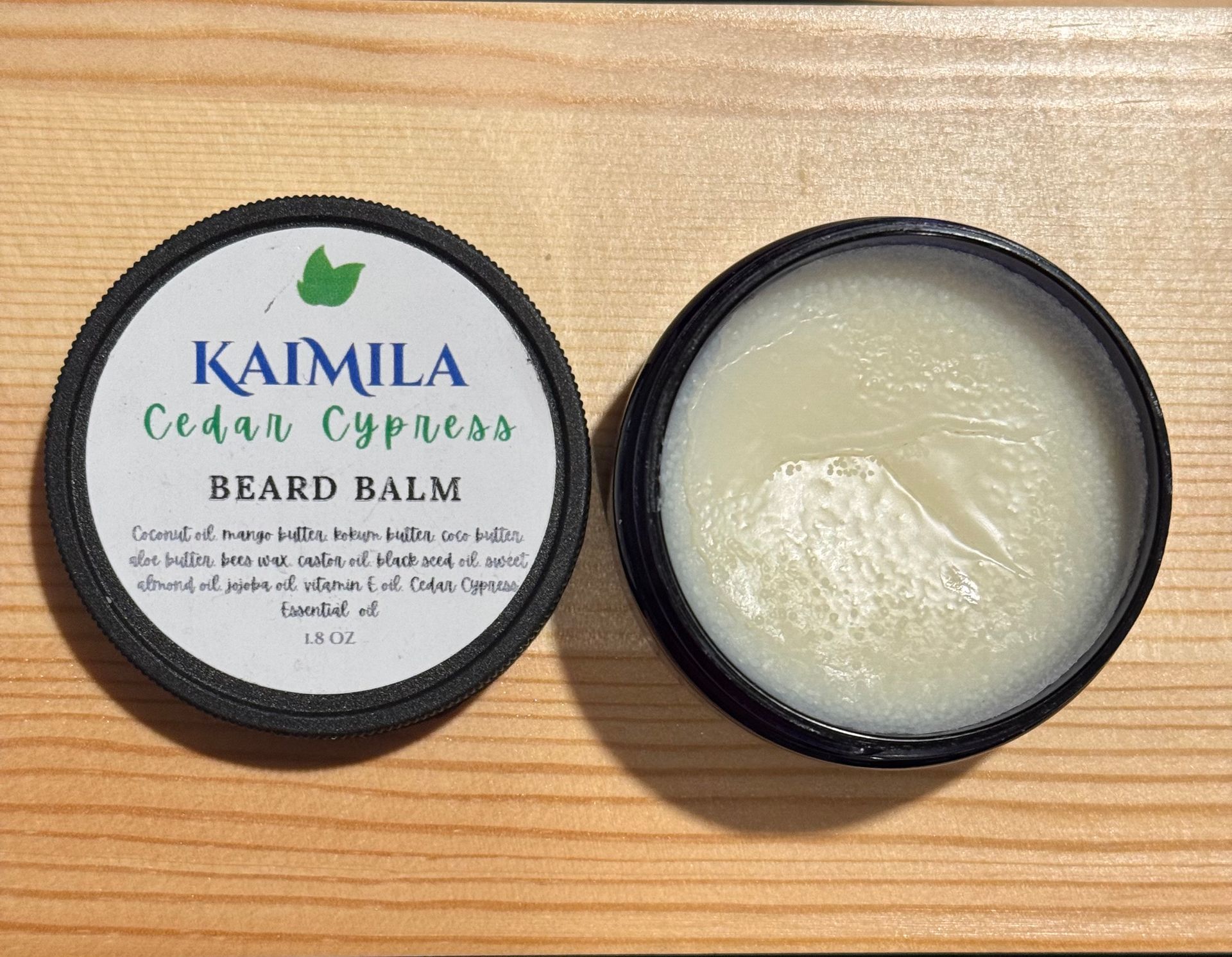 Black jar of beard balm, with label “Kaimila Cedar Cypress Beard Balm” next to its open lid.