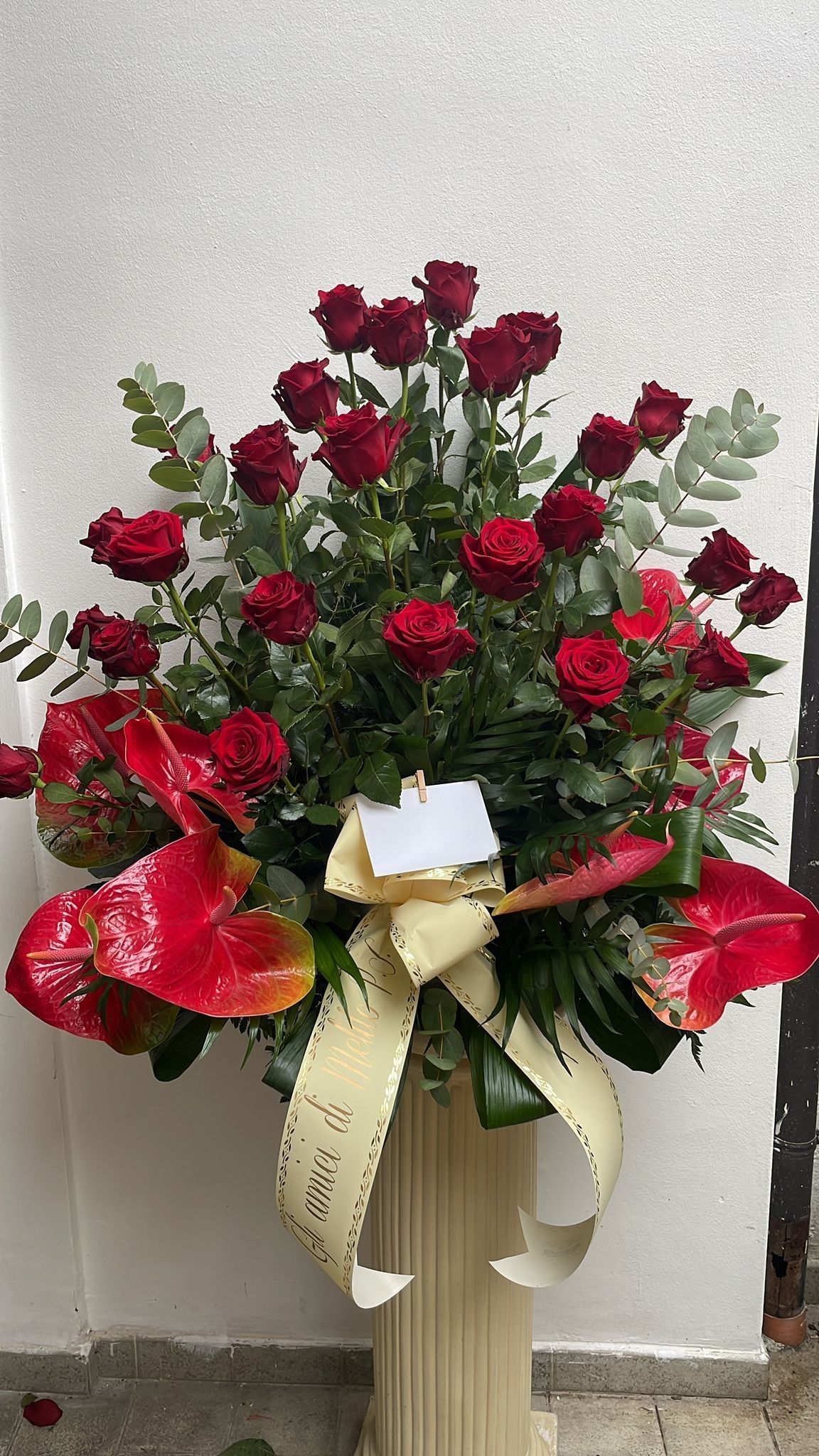 Composizione floreale di rose rosse e anthurium con nastro color crema in un vaso color crema, contro una parete bianca testurizzata.