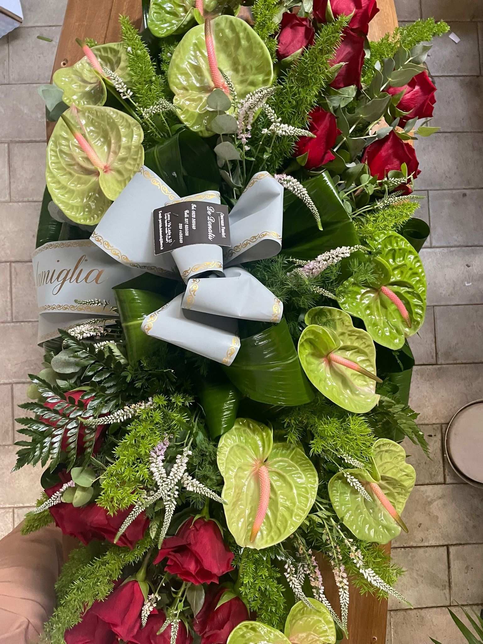 Composizione floreale con rose rosse, anthurium verdi e un fiocco azzurro.