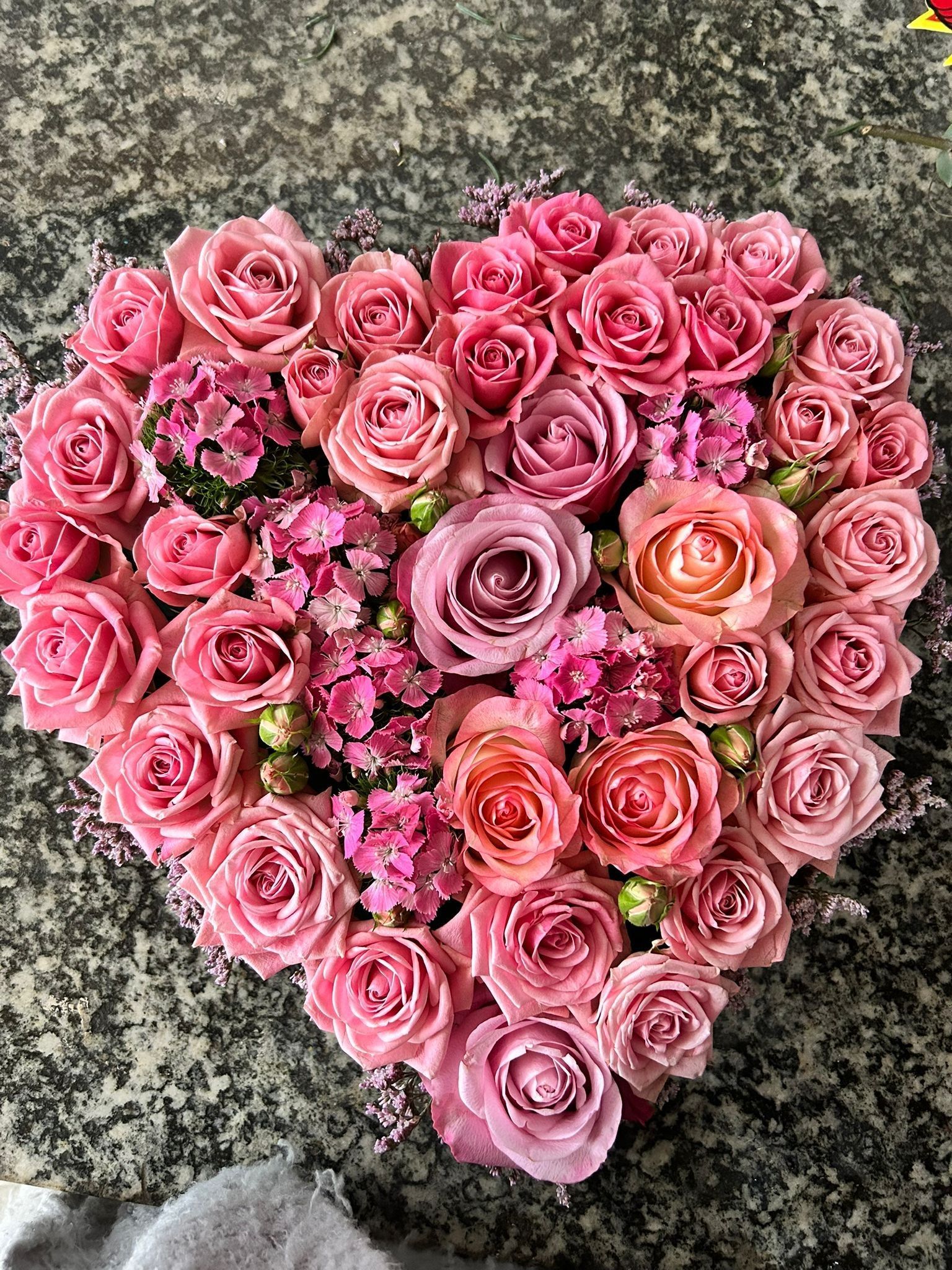Composizione floreale a forma di cuore composta da rose rosa e altri fiori rosa su una superficie strutturata.
