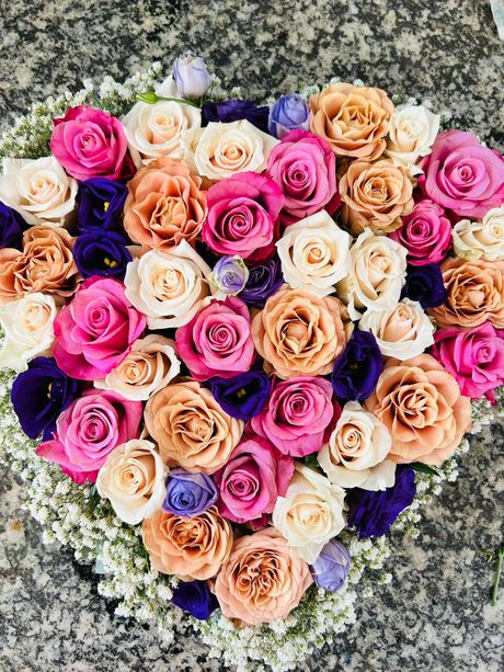 Composizione floreale a forma di cuore con rose rosa, pesca, avorio e accenti viola, appoggiata su gypsophila bianca.