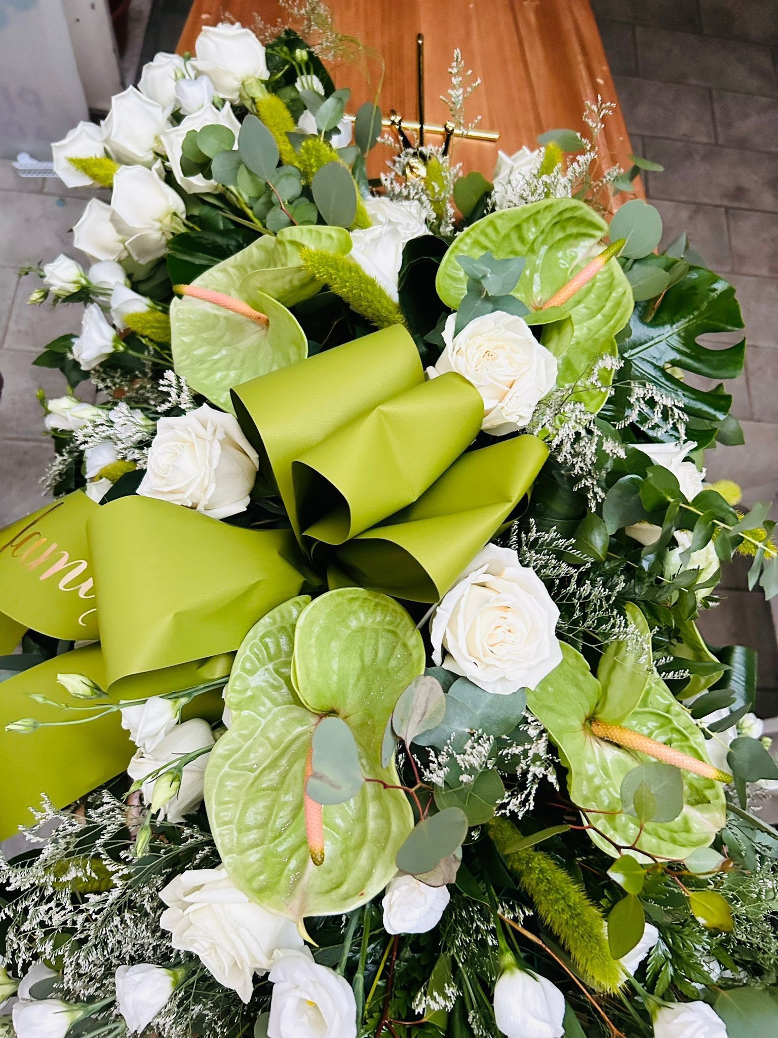 Composizione floreale funebre su una bara, con rose bianche, anthurium e dettagli verdi.