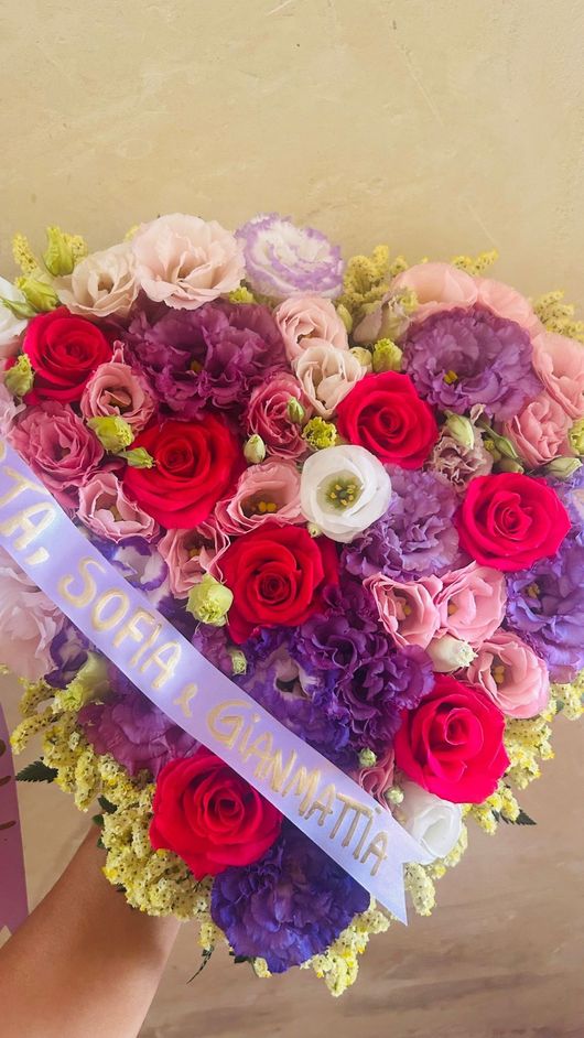 Composizione floreale a forma di cuore con fiori rossi, viola, rosa e bianchi, con un nastro con la scritta 