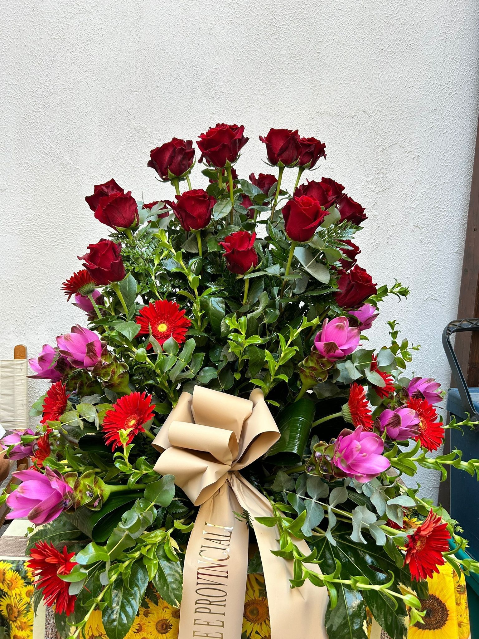 Composizione floreale con rose rosse, fiori rosa e rossi e un nastro beige.