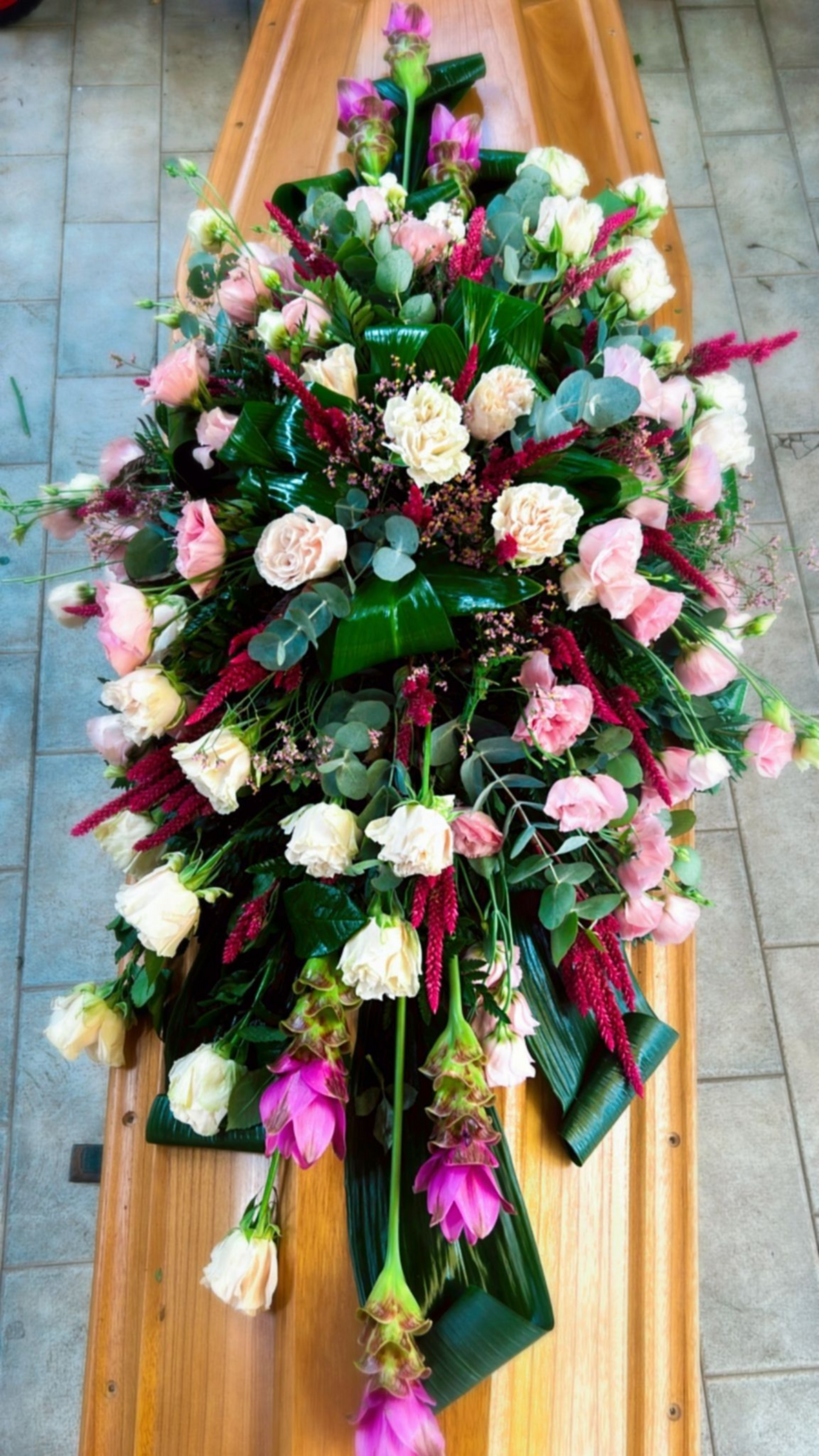 Composizione floreale nei toni del rosa, del bianco e del rosso, adagiata su una bara di legno.