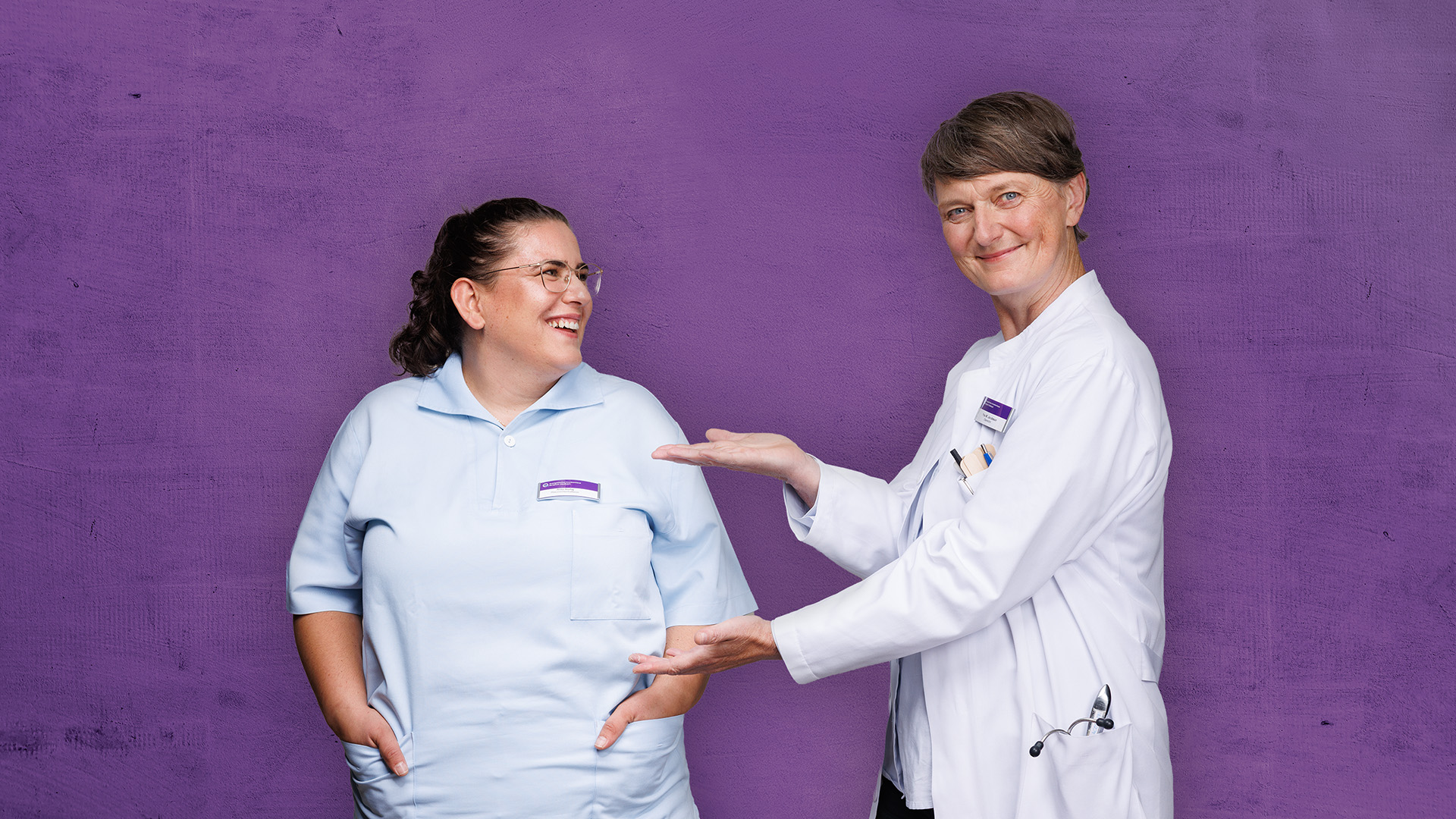 Zwei Personen in weißen Kitteln lächeln und gestikulieren vor einem violetten Hintergrund.