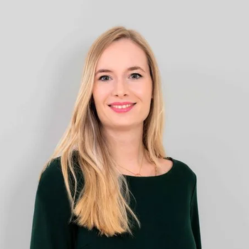 Lächelnde blonde Frau in einem dunkelgrünen Oberteil vor hellgrauem Hintergrund