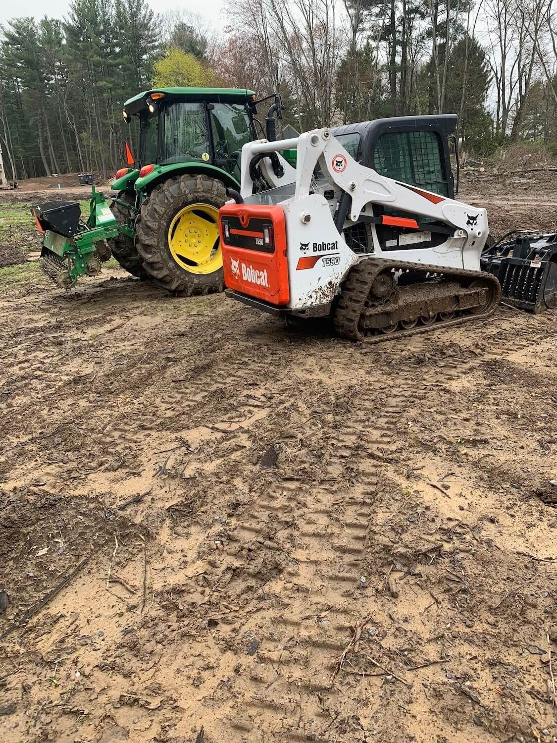 A green John Deere tractor and a white Bobcat compact track loader parked on a muddy, cleared construction site.
