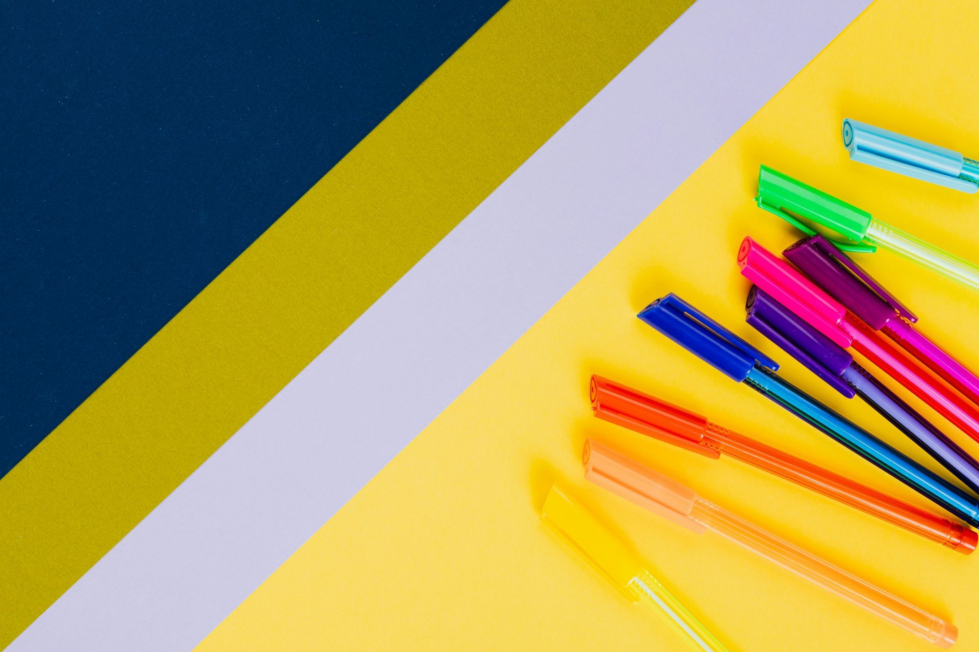 Bolígrafos de colores sobre fondo amarillo con bloques de color en ángulo: azul marino, dorado y lavanda.