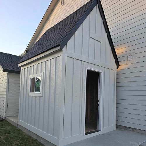Exterior of a small white shed-like structure attached to a house. It has a window, a dark door, and a black roof.