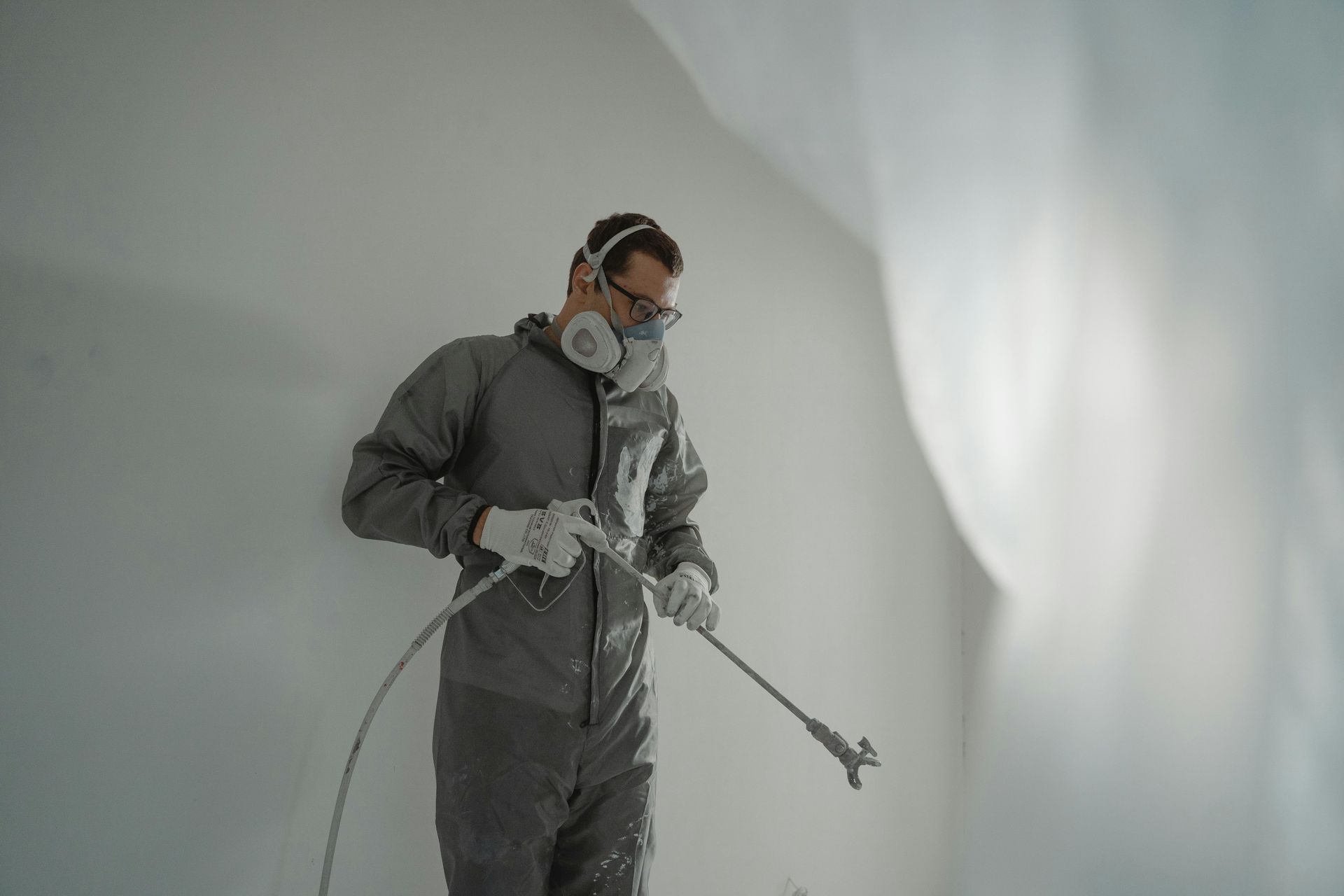 Person in protective suit, mask, and gloves spraying white paint on a wall.