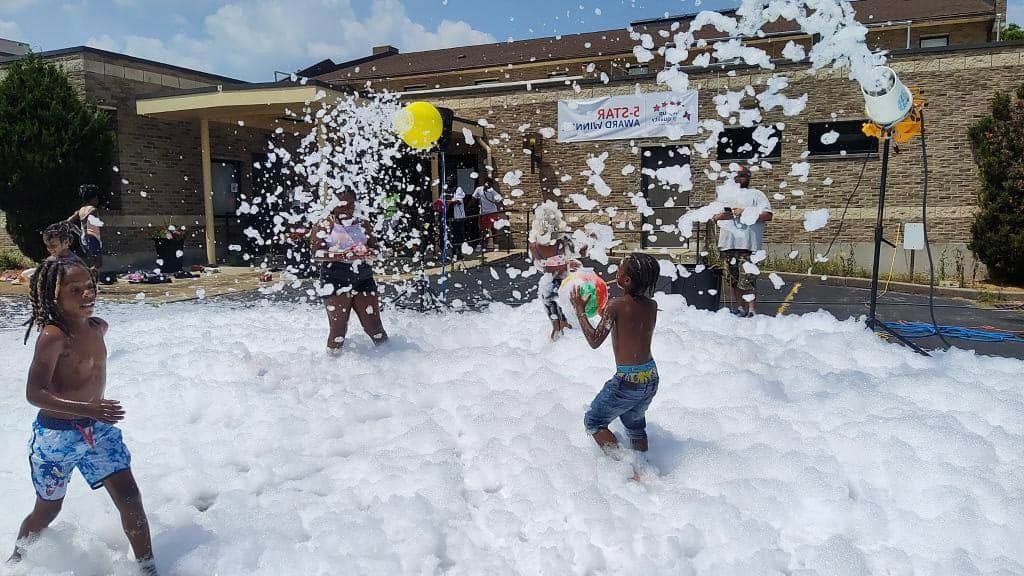A group of children are playing in foam in front of a building.
