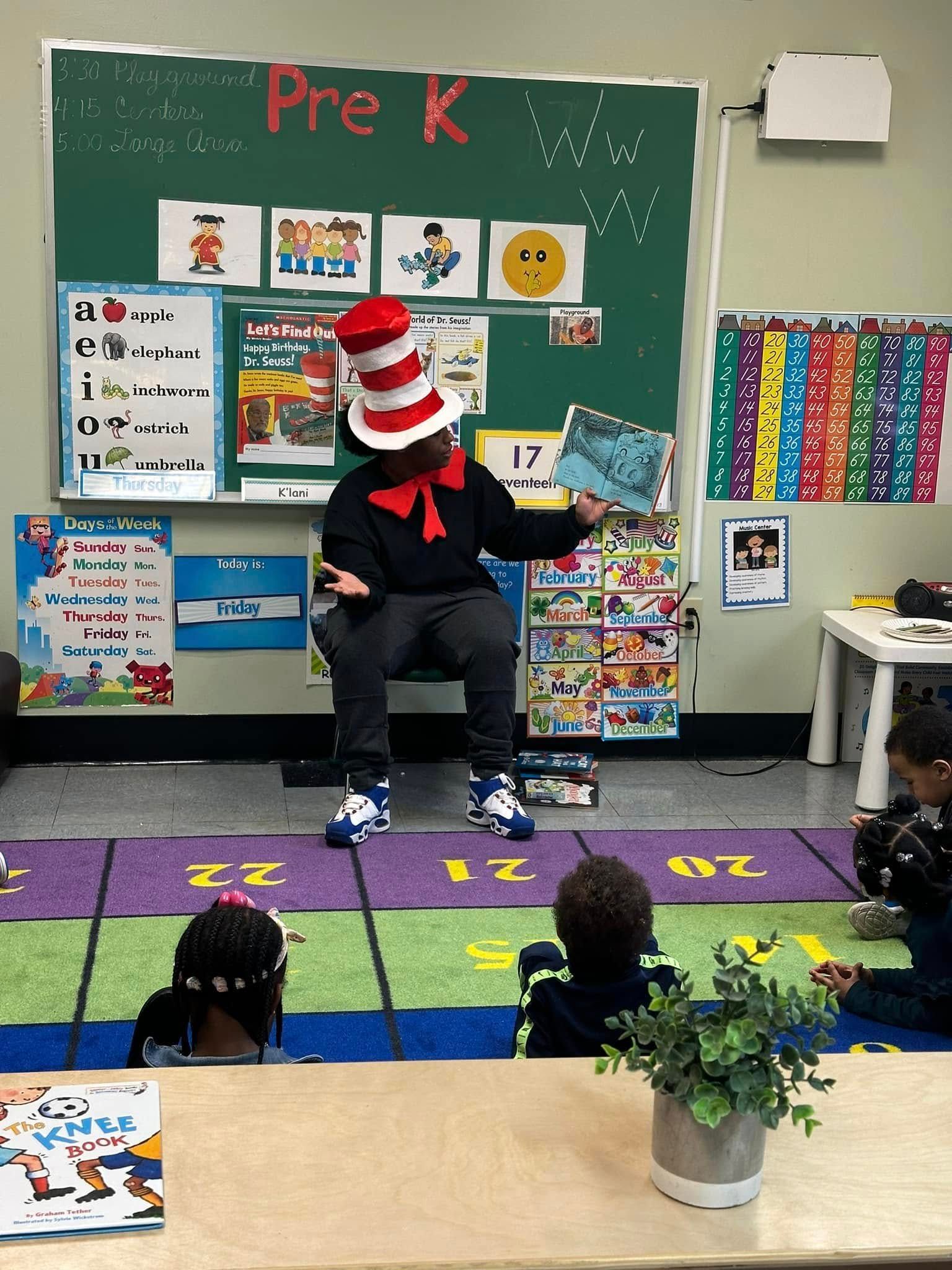 A man in a cat in the hat outfit is reading a book to children in a classroom.