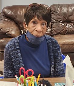A woman wearing a mask is sitting at a table with scissors and pencils.