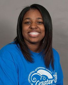 A woman wearing a blue shirt with the word greater on it is smiling.