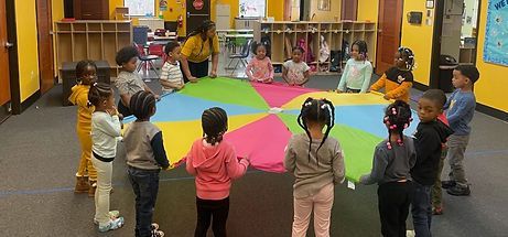 A group of children are standing around a colorful parachute in a classroom.