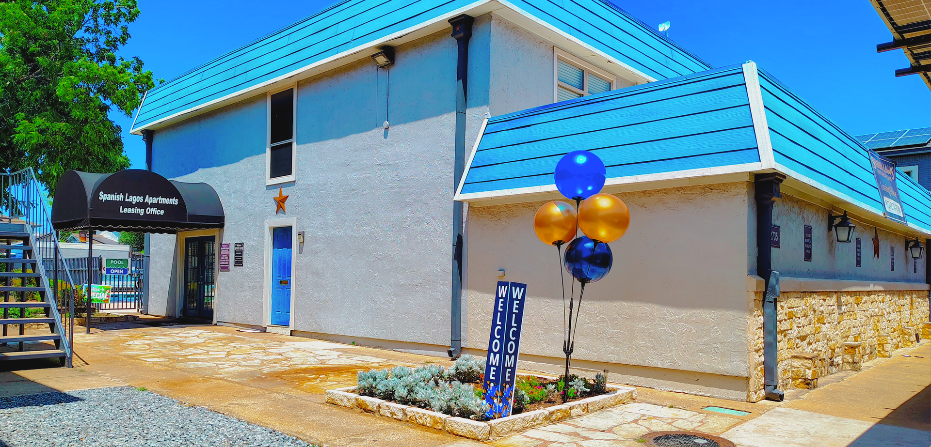 Exterior of a light blue building with a turquoise roof. Balloons and a sign are in front of the building, with a tree on the left side.