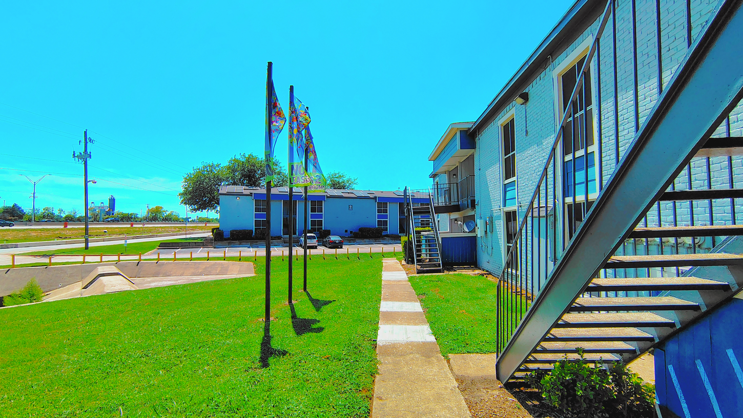 Blue apartment building with grassy yard, walkway, and flagpoles under a clear blue sky.