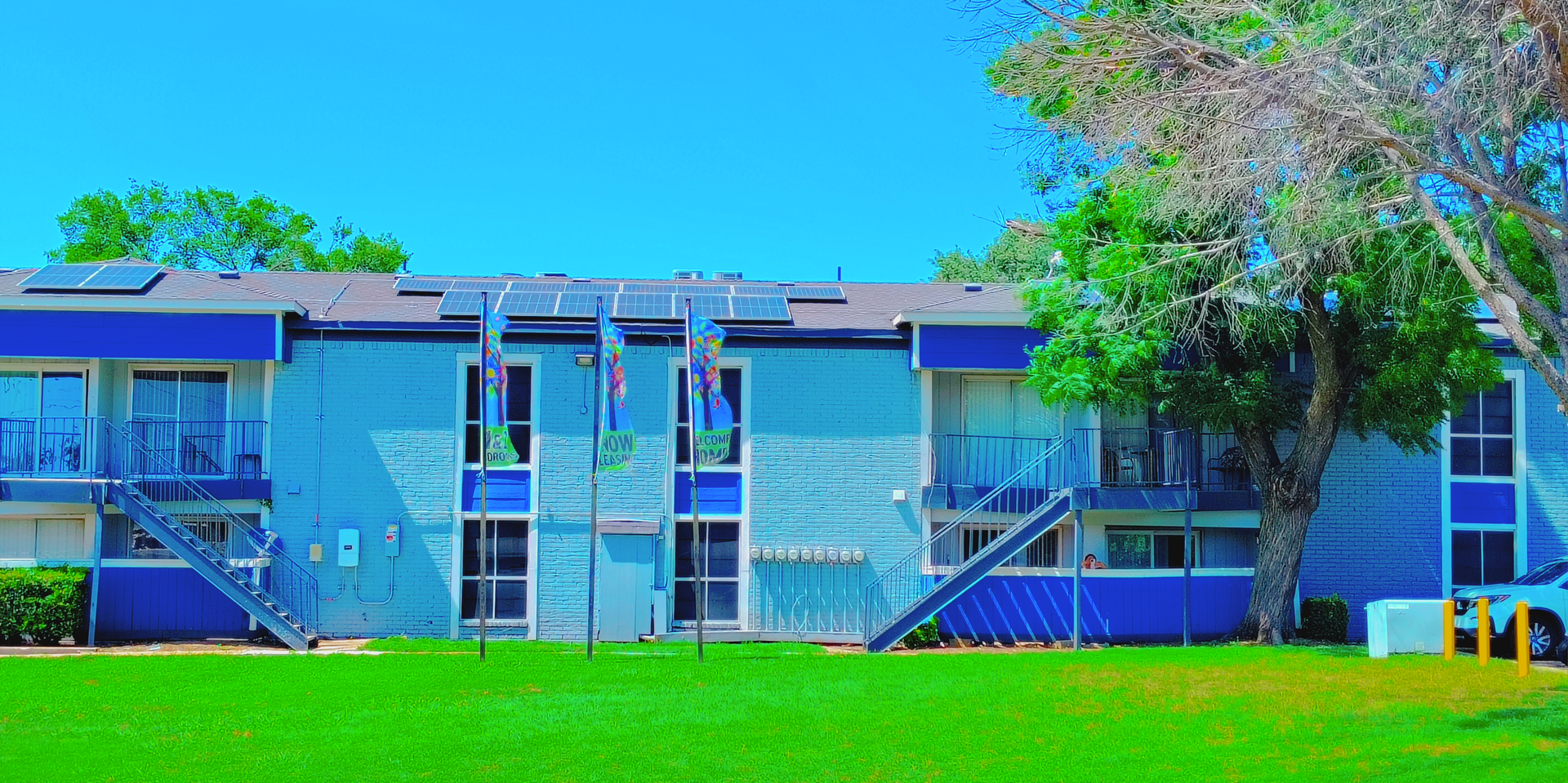 Two-story blue apartment building with dark blue trim, solar panels on the roof, and green lawn. The building has a blue sky background and a large tree on the right side.