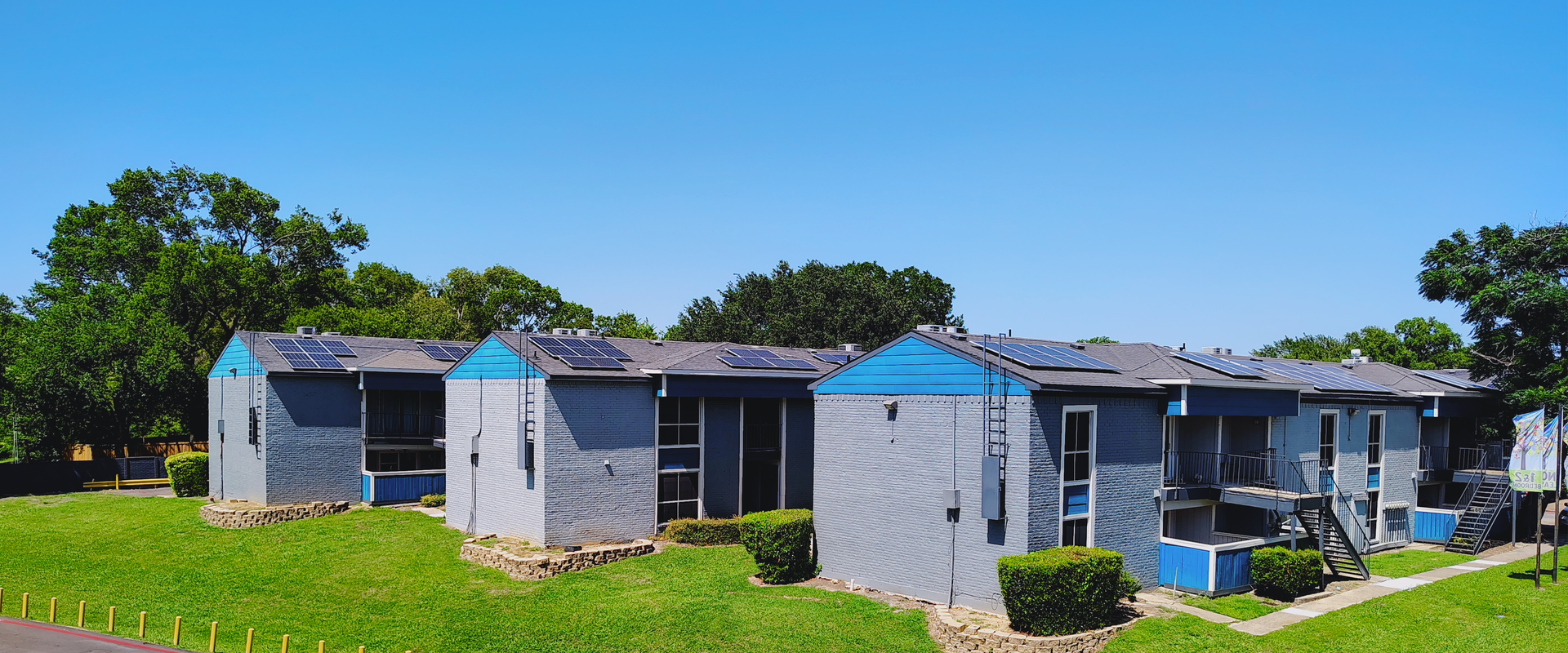Row of small, blue-roofed buildings with solar panels on the roofs. Green grass and trees are in the foreground under a blue sky.