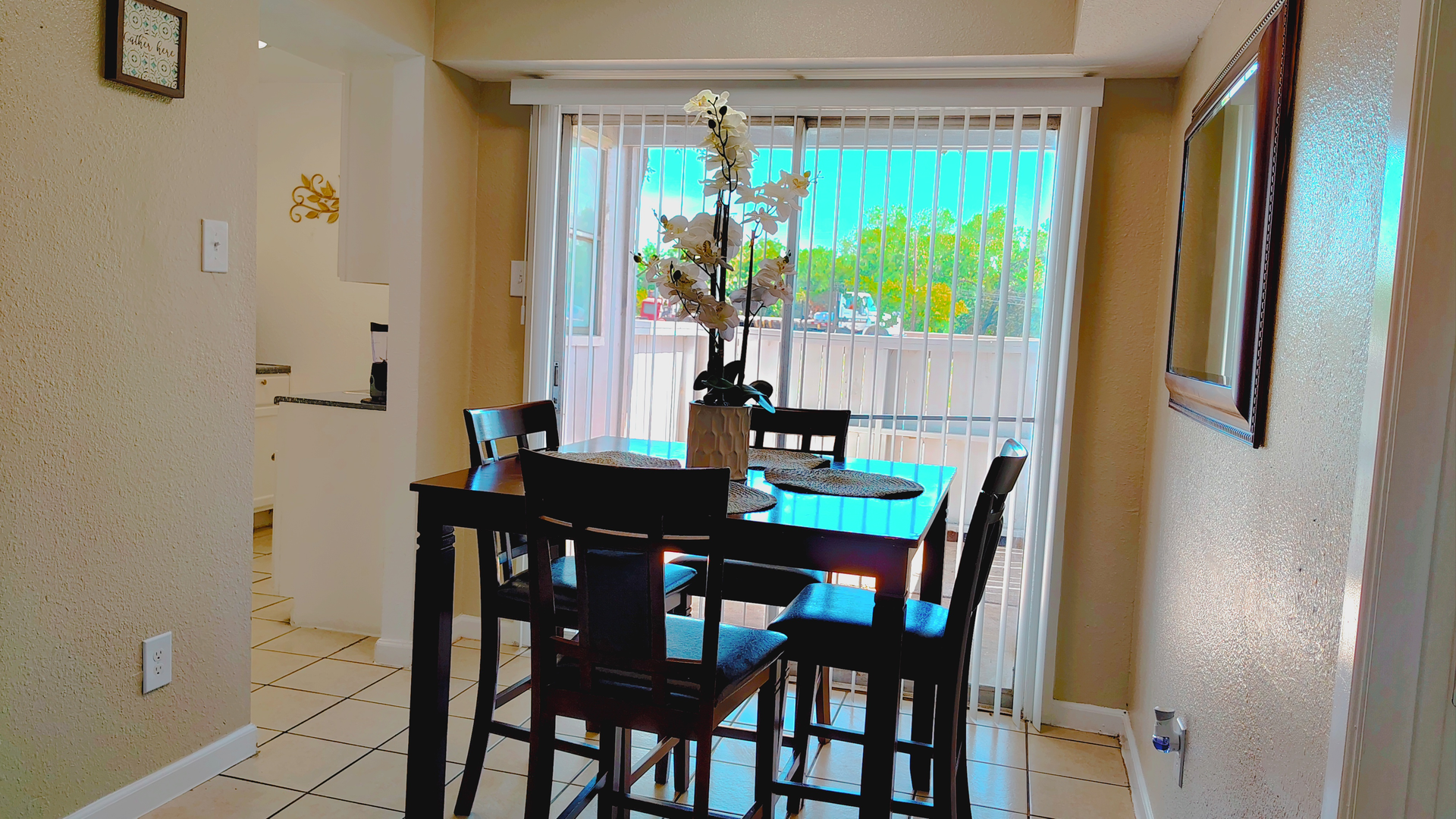 Dining area with a dark wood table and chairs, a vase of flowers, and a sliding glass door with a view of trees.