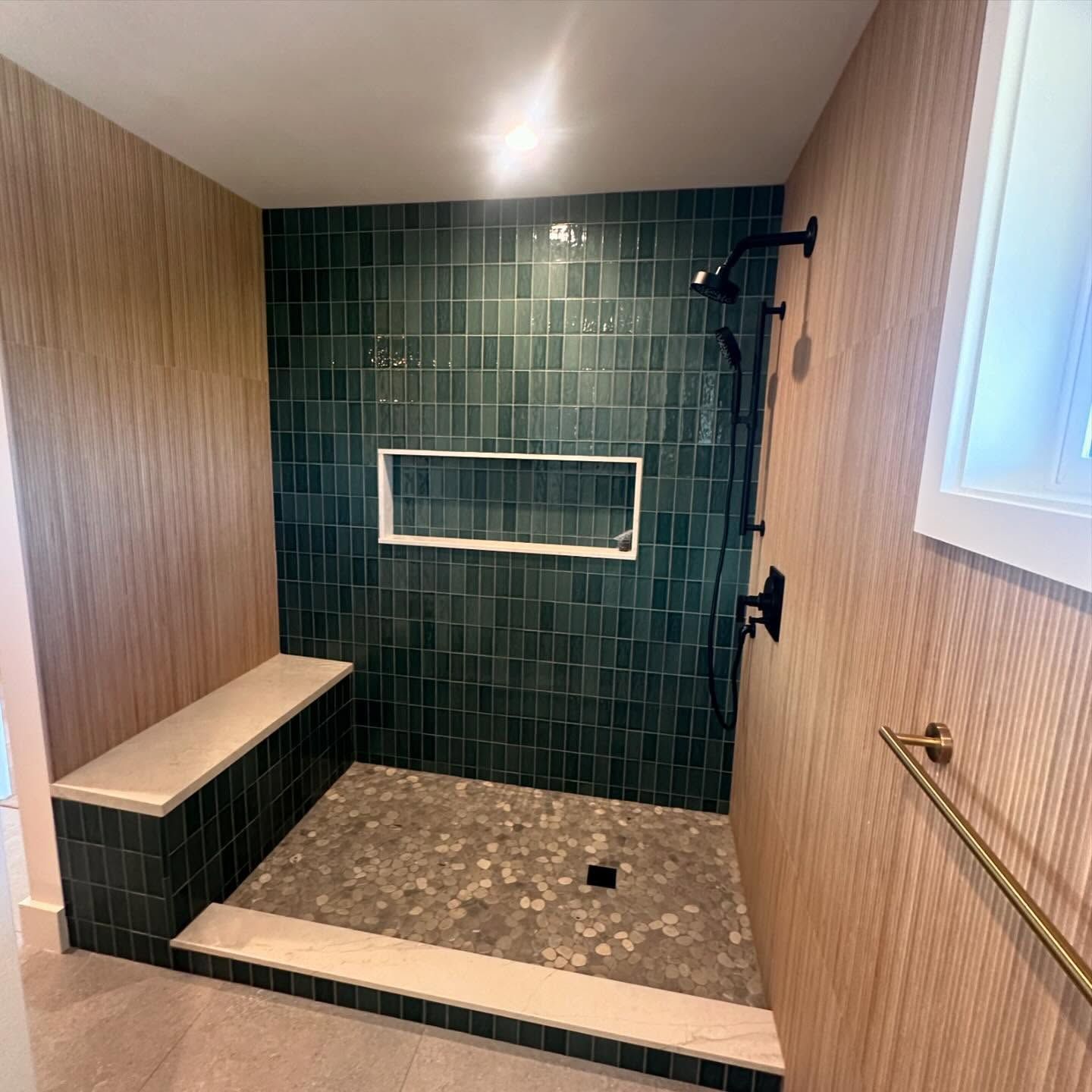 Shower with green tile wall and stone floor, bench. Wood panel walls. Black shower fixtures.