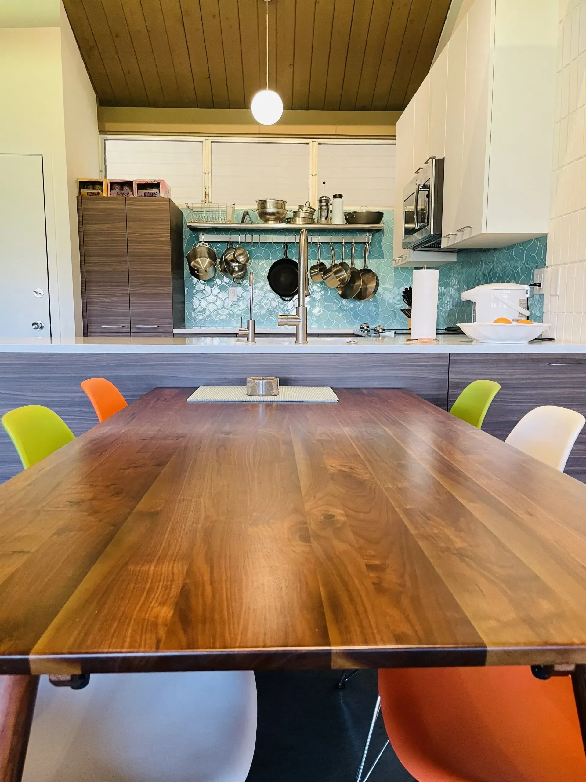 A long wooden dining table with colorful chairs, in front of a kitchen with blue tile.
