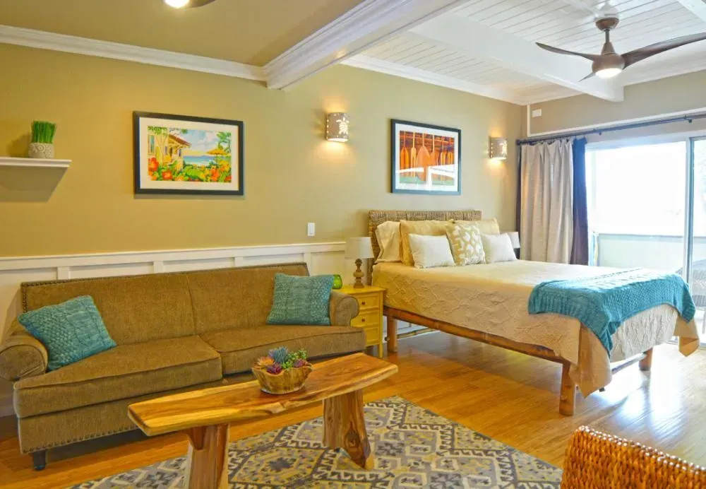 Cozy studio apartment with a bed, couch, and balcony, light tan walls, and wooden floors.