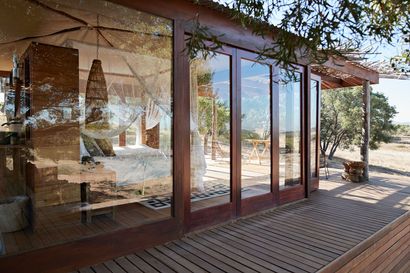 Wooden cabin with glass doors. Bedroom with canopy bed visible. Porch with wooden deck and surrounding nature.
