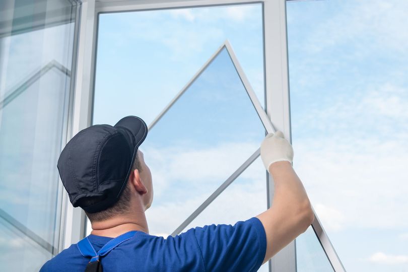 Man in blue shirt and cap installing a screen window, outdoors, against a blue sky.