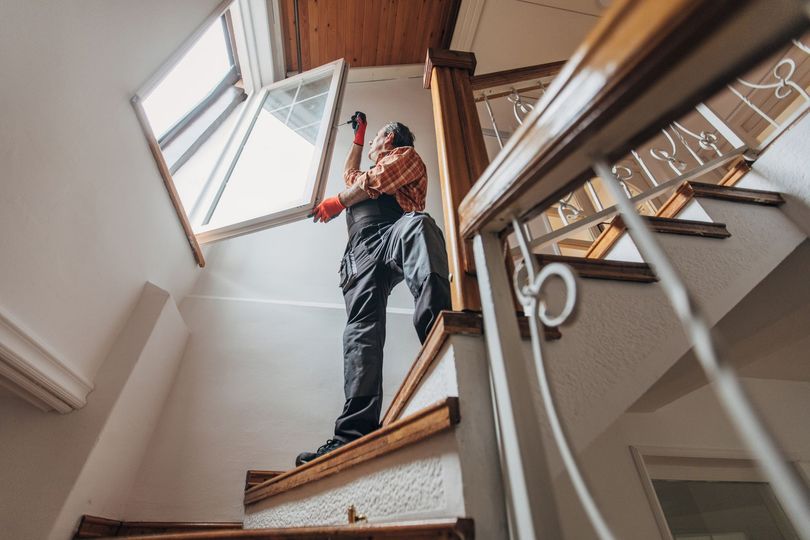 Man in work clothes on stairs holding a window pane, preparing to install it.