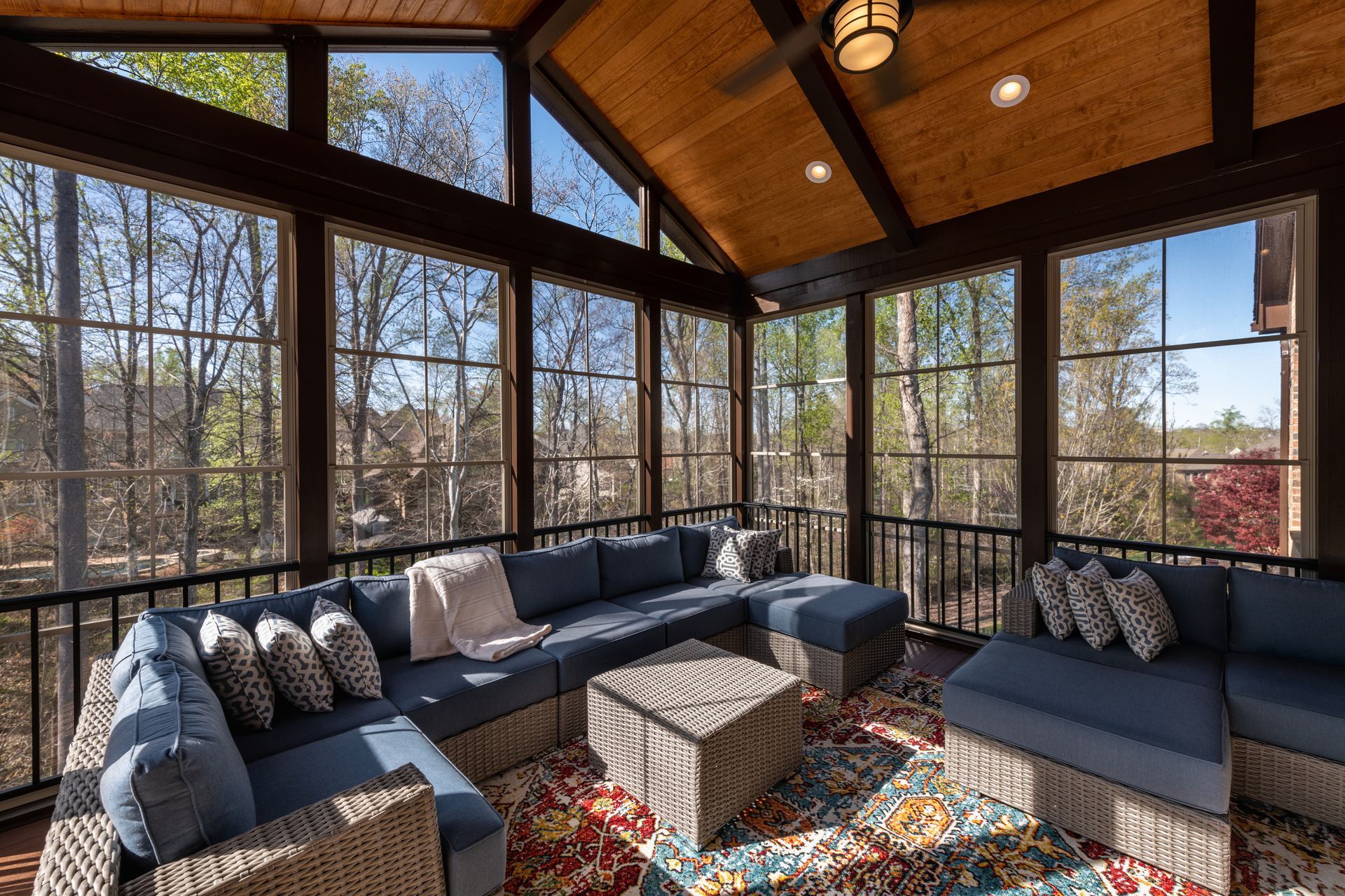Sunroom with blue sectional sofa, patterned rug, and large windows overlooking trees.