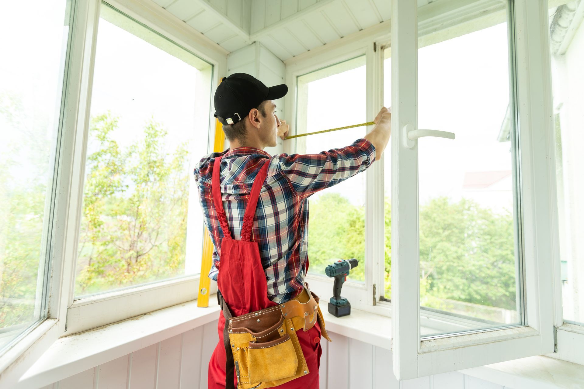 A man in red overalls measures a window frame with a measuring tape.